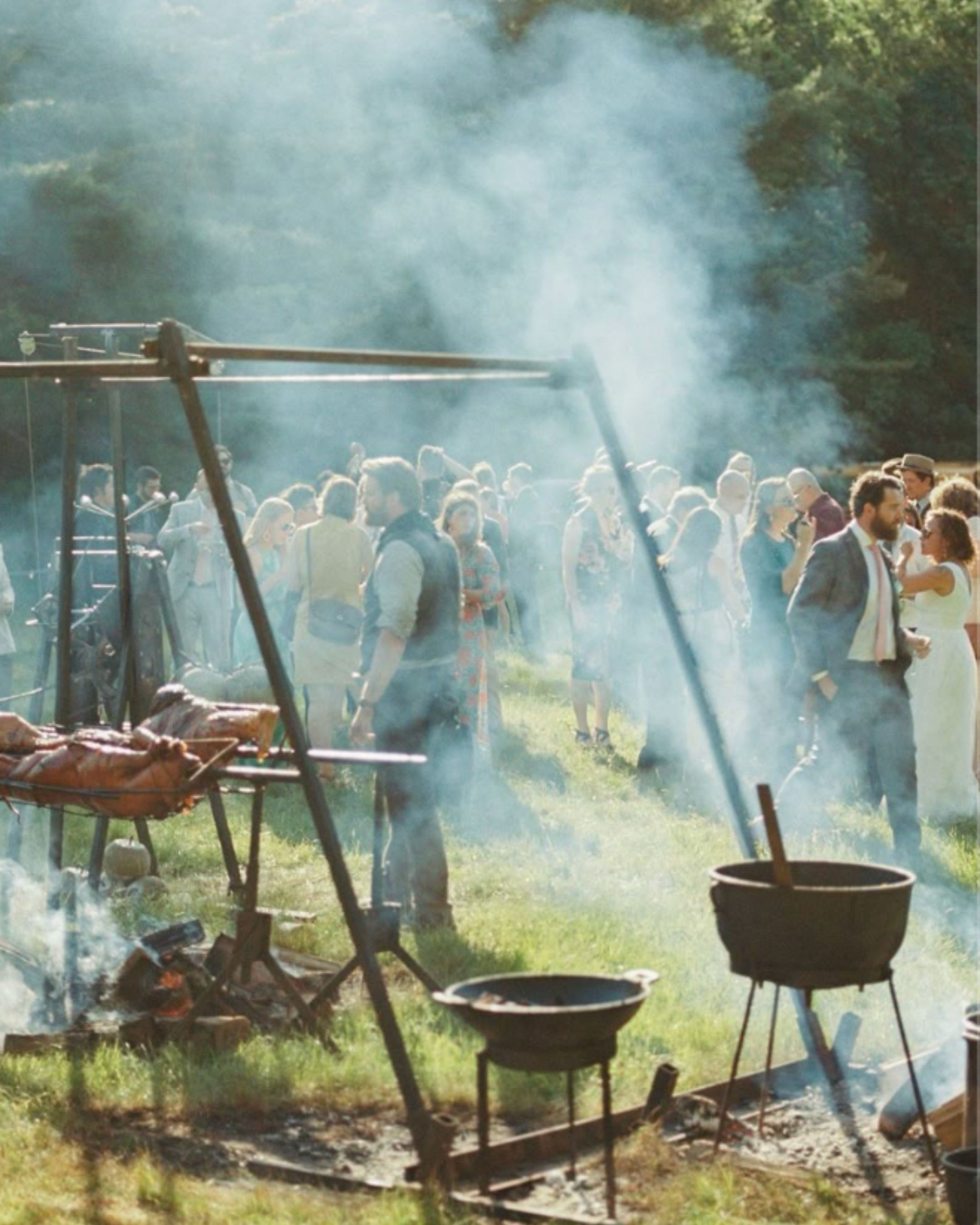 Barbecue grill with food and smoke, outdoor setting with trees and people in the background.