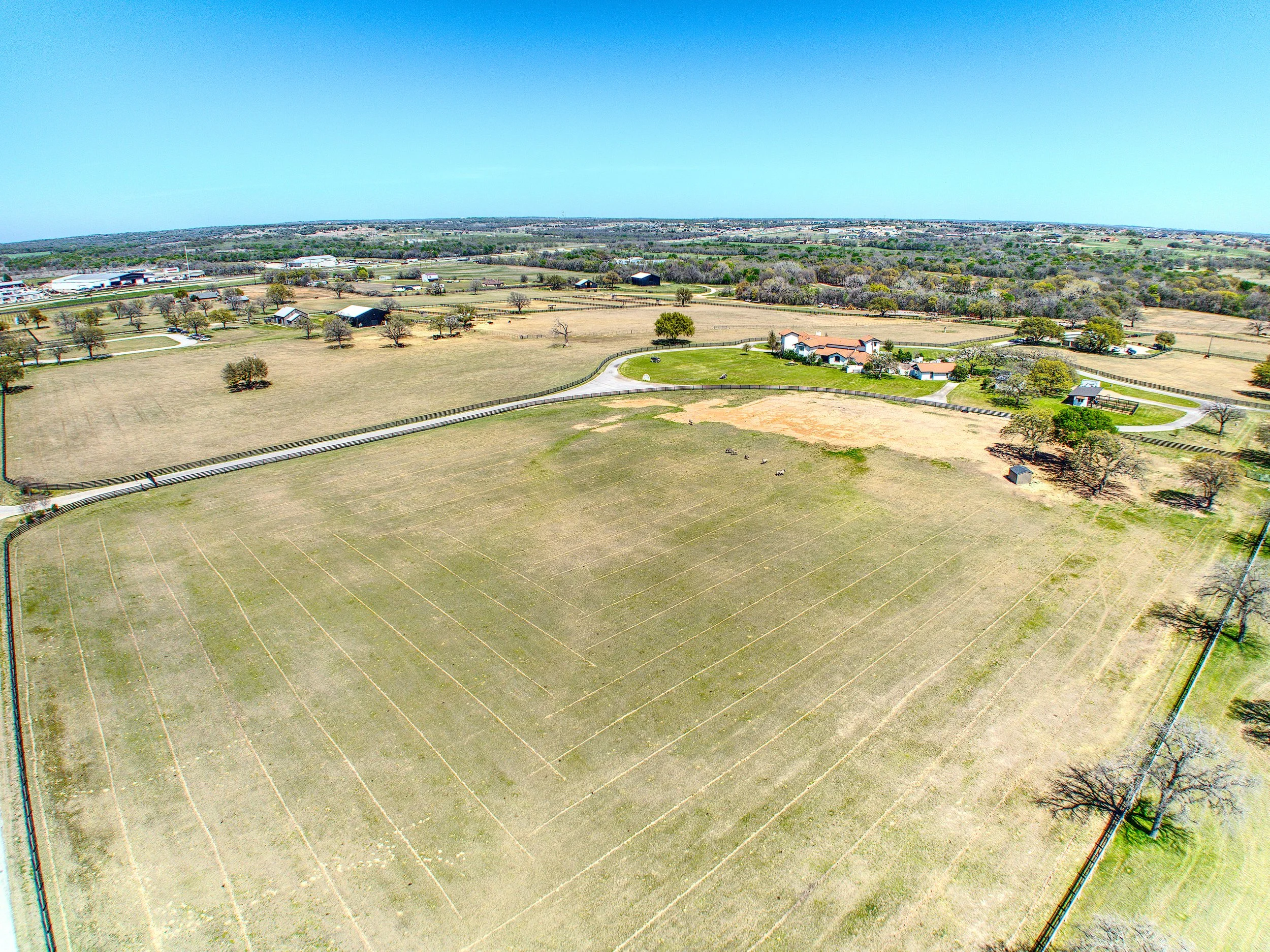 Aerial view of a rural farmland with trees, fields, and small buildings under a clear blue sky.