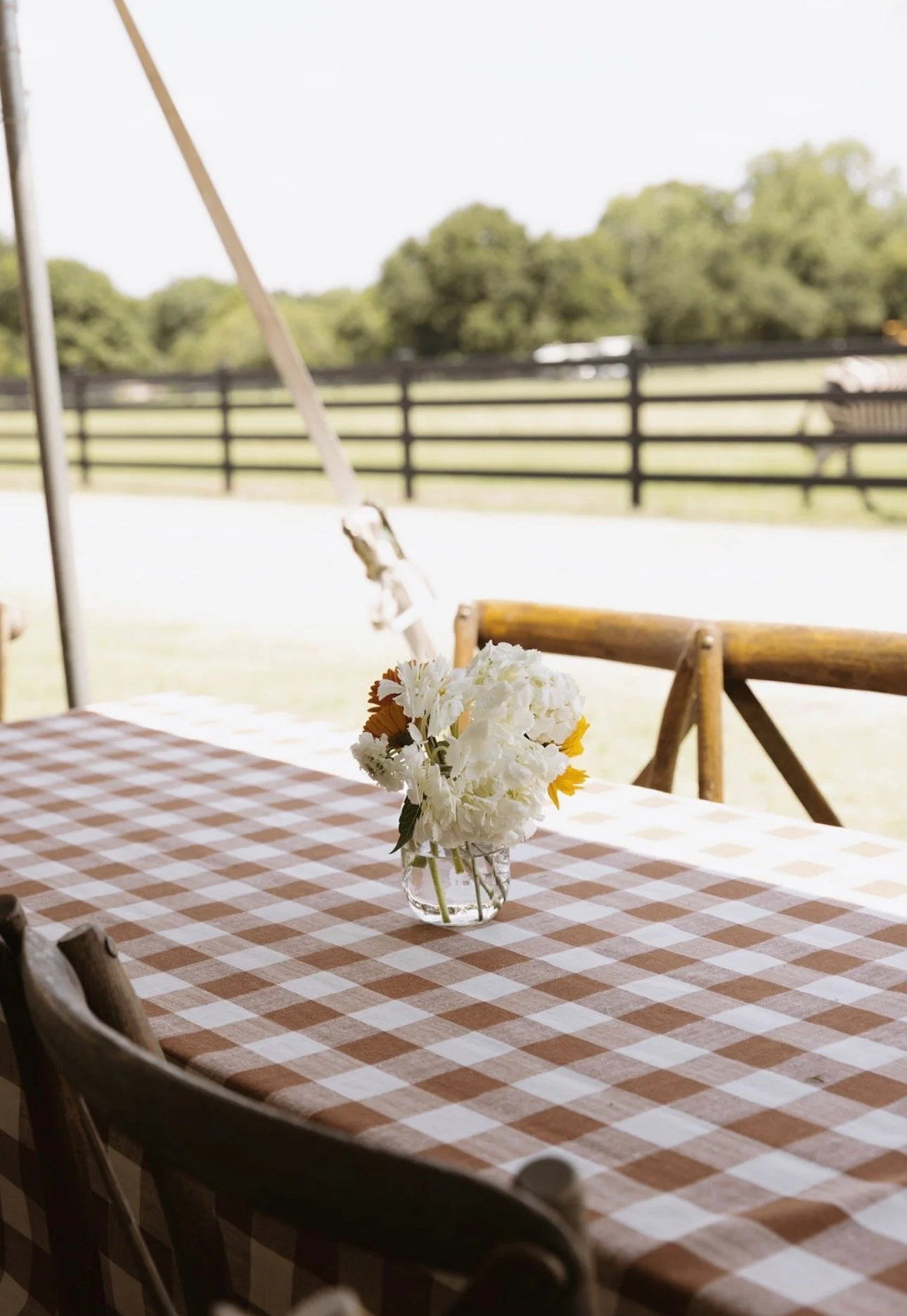 A table with a checkered tablecloth and a small glass vase holding white and yellow flowers, set outdoors with a wooden fence and green trees in the background.