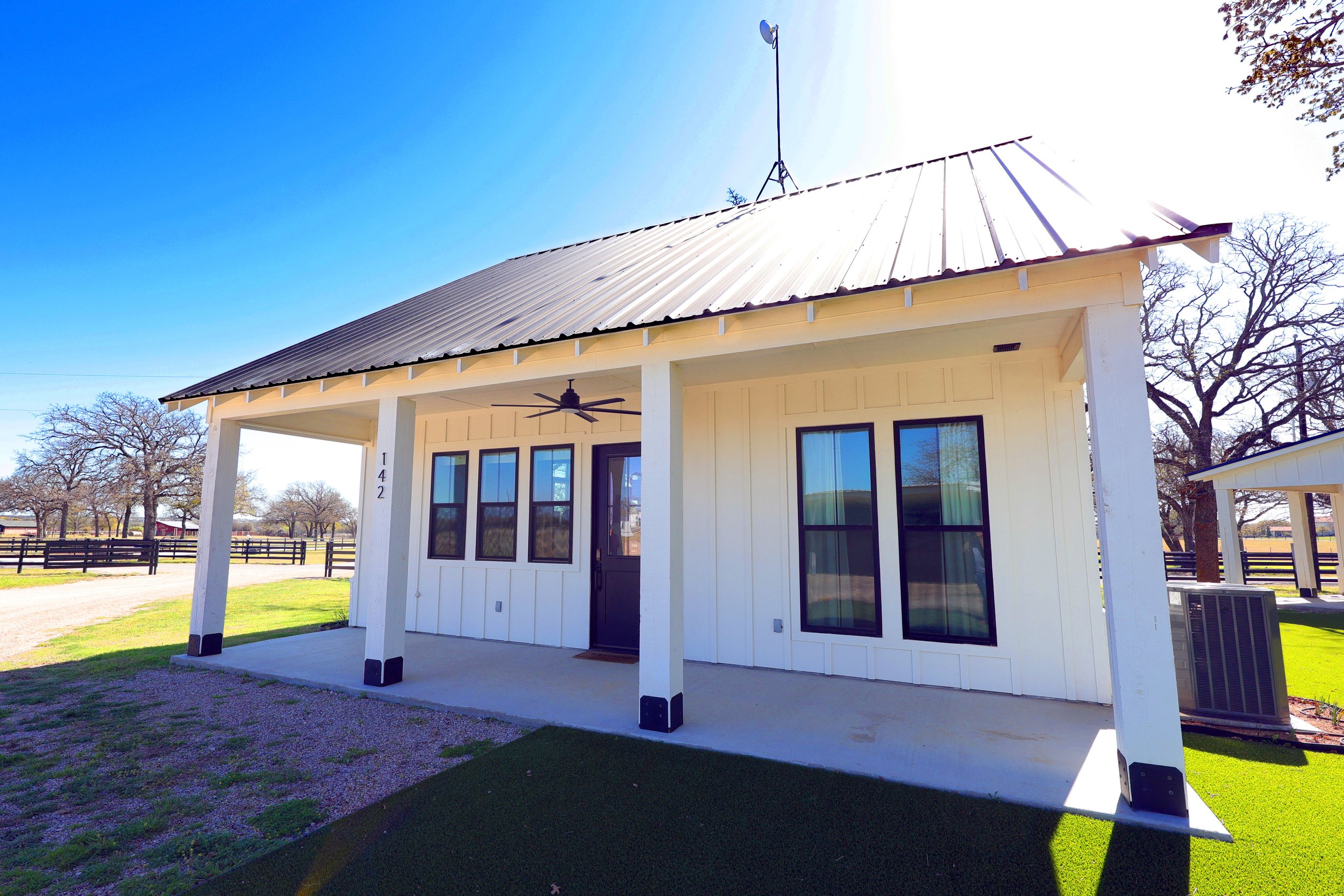 A white house with a metal roof, large windows, and a covered porch, situated in a rural area with trees and open land in the background under a clear blue sky.