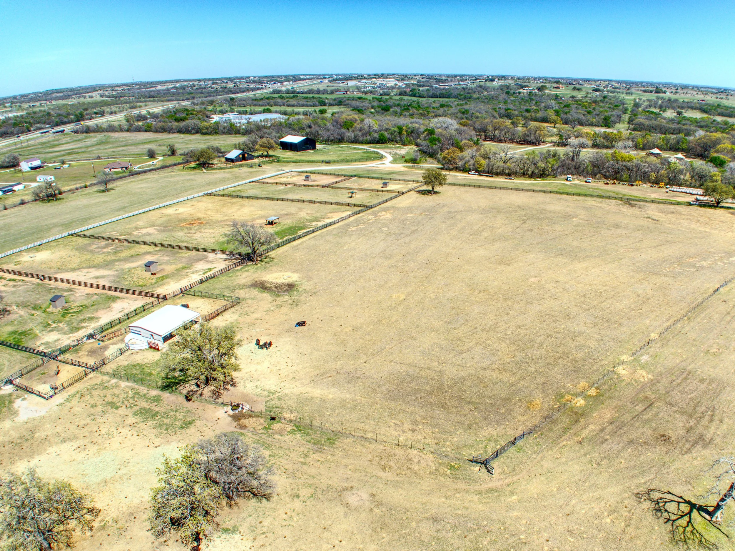 Aerial view of farmland with fenced areas, barns, trees, and a few horses, with a distant view of a city and roads under a clear blue sky.