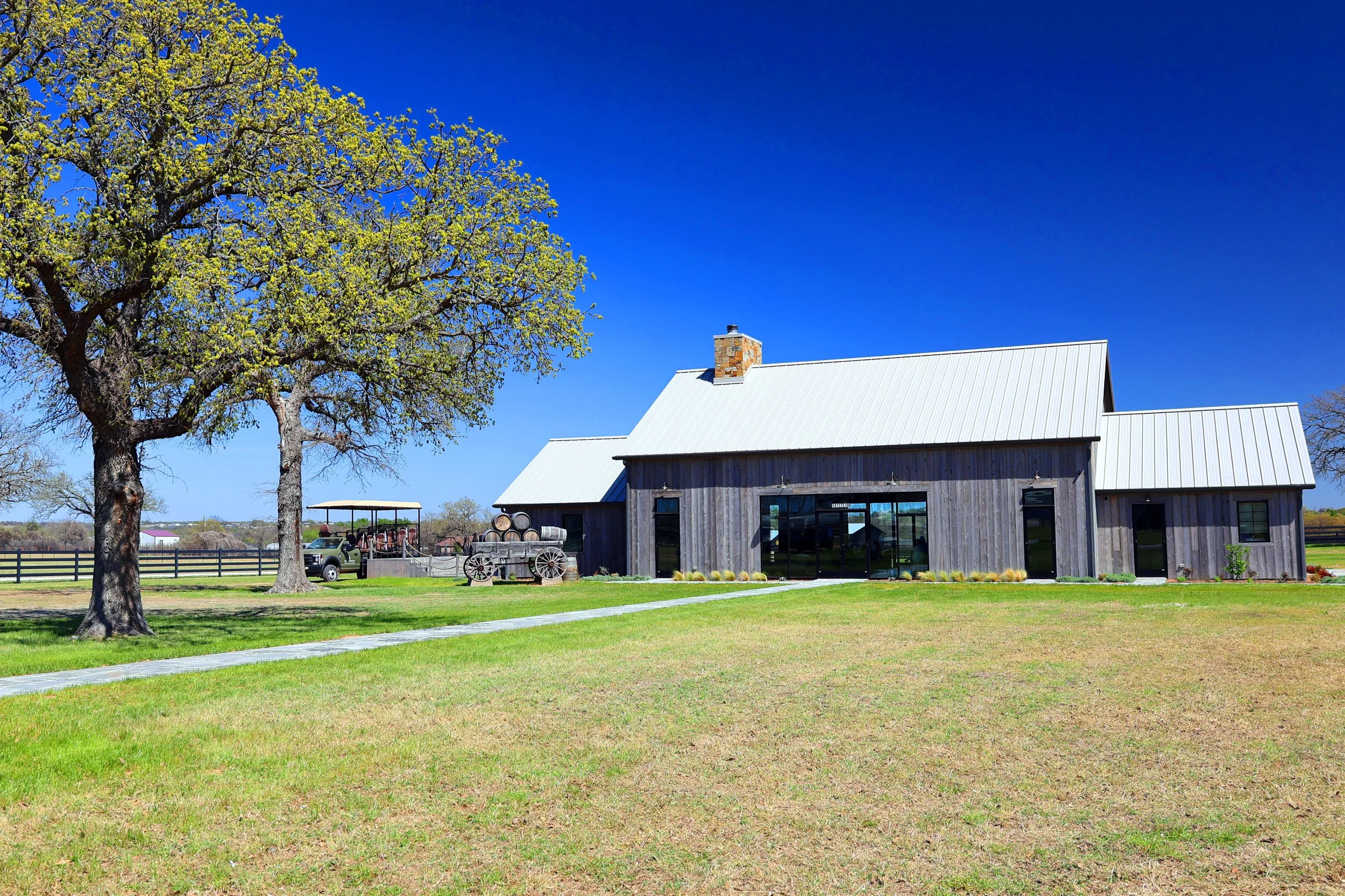 A rural landscape featuring a wooden barn with a metal roof, a large tree with budding green leaves, and a tractor with a wagon in the background under a bright blue sky.