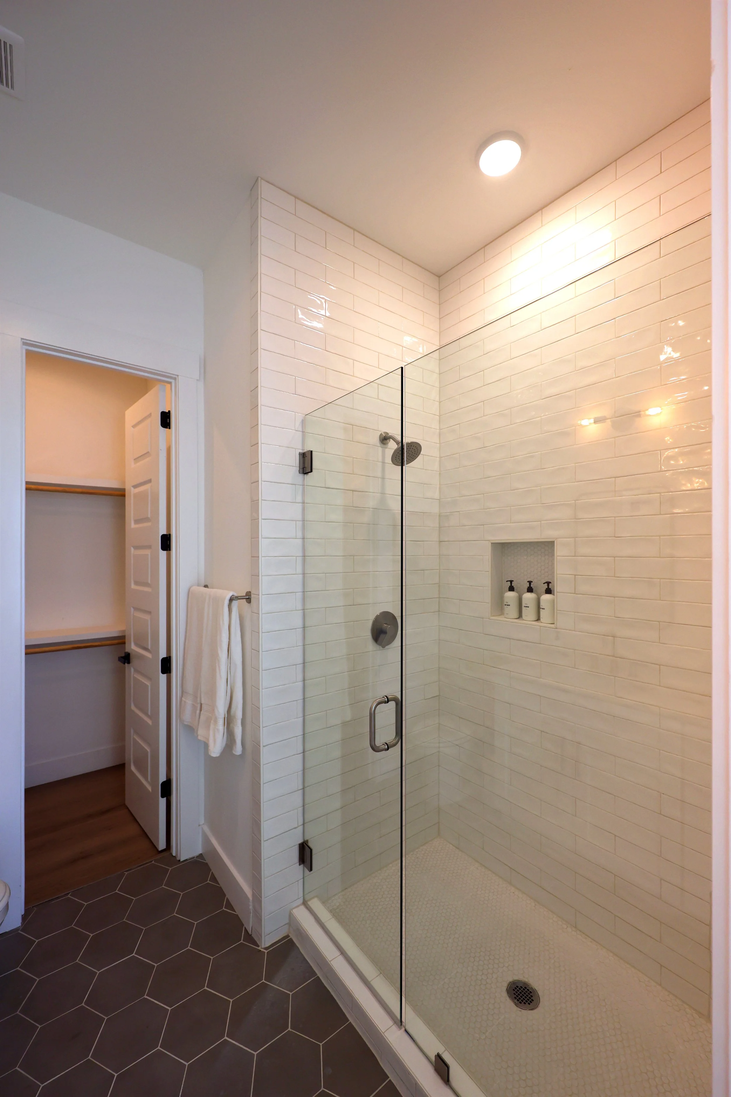A modern walk-in shower with glass enclosure, white subway tile walls, and a built-in shower niche with three bottles. The shower has a rainfall showerhead and a drain on the textured white floor. Adjacent is a doorway leading to a walk-in closet wit