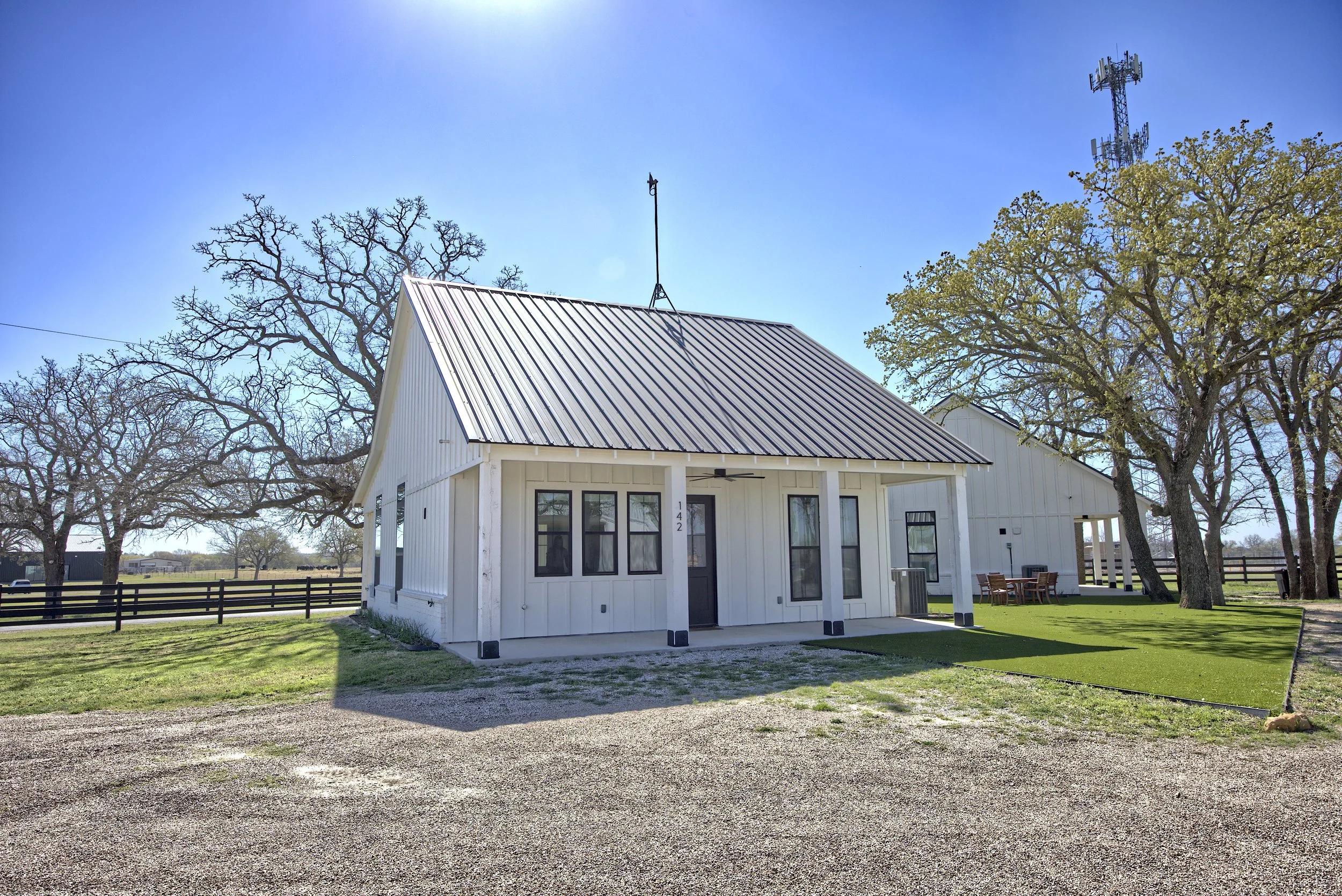 White modern house with metal roof, surrounded by green lawn, trees, and fencing under a clear blue sky.