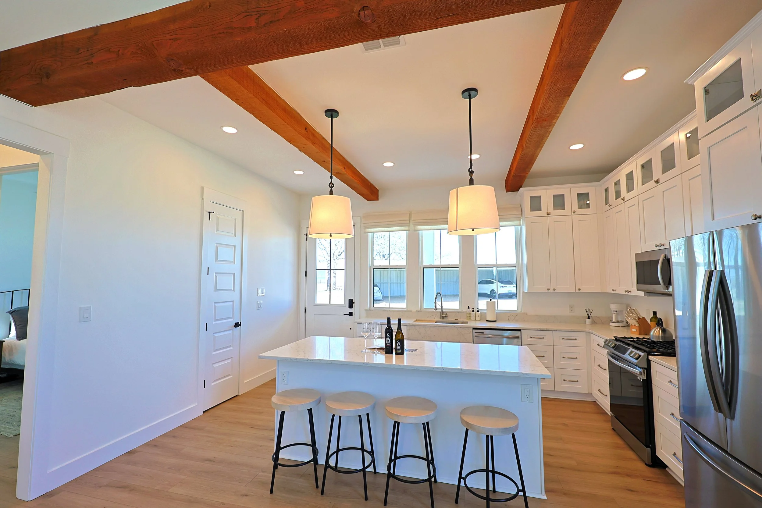 A modern kitchen with a white countertop island, four stools, and white cabinets. Two pendant lights hang above the island, and natural light comes through large windows. There are wine bottles and glasses on the island.