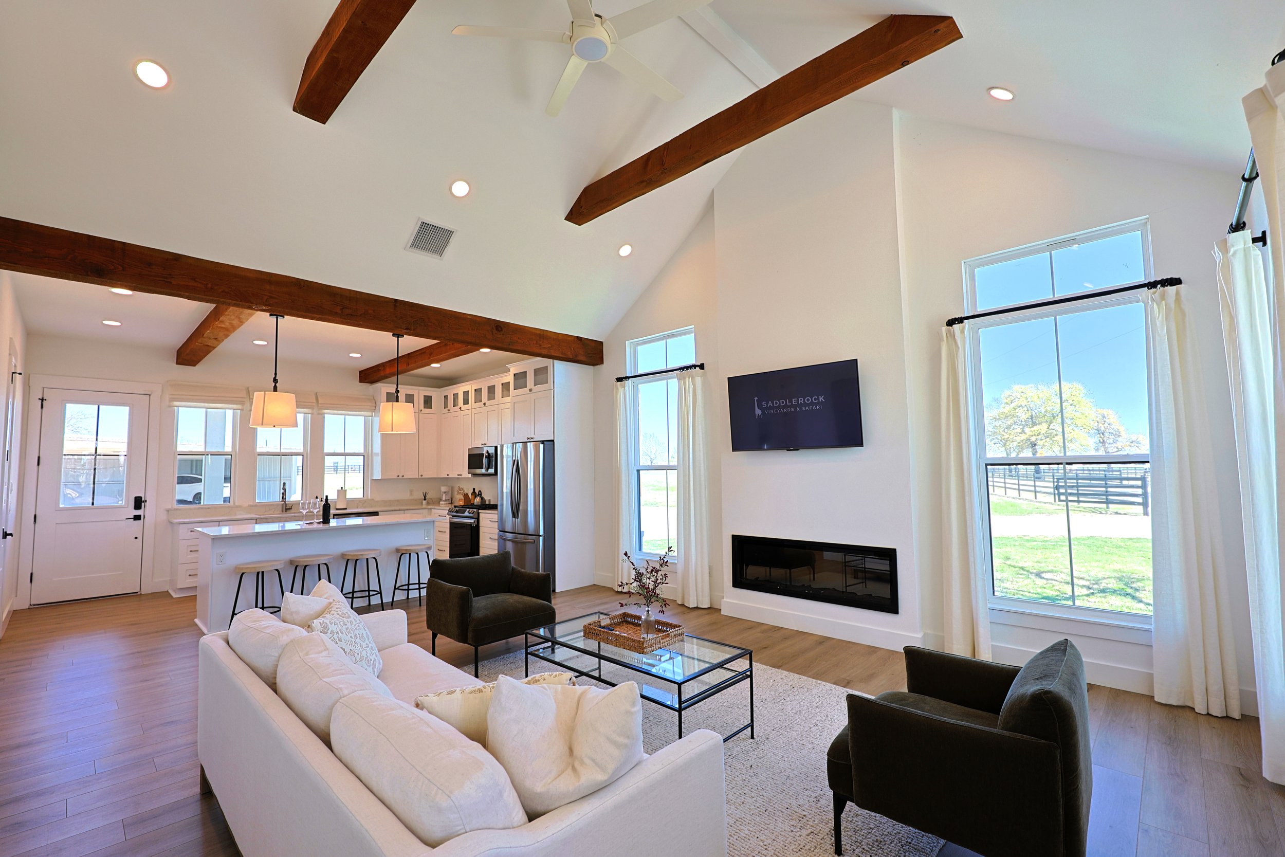 Open-concept living room and kitchen with vaulted ceiling, wooden beams, large windows with curtains, white walls, and modern furniture.