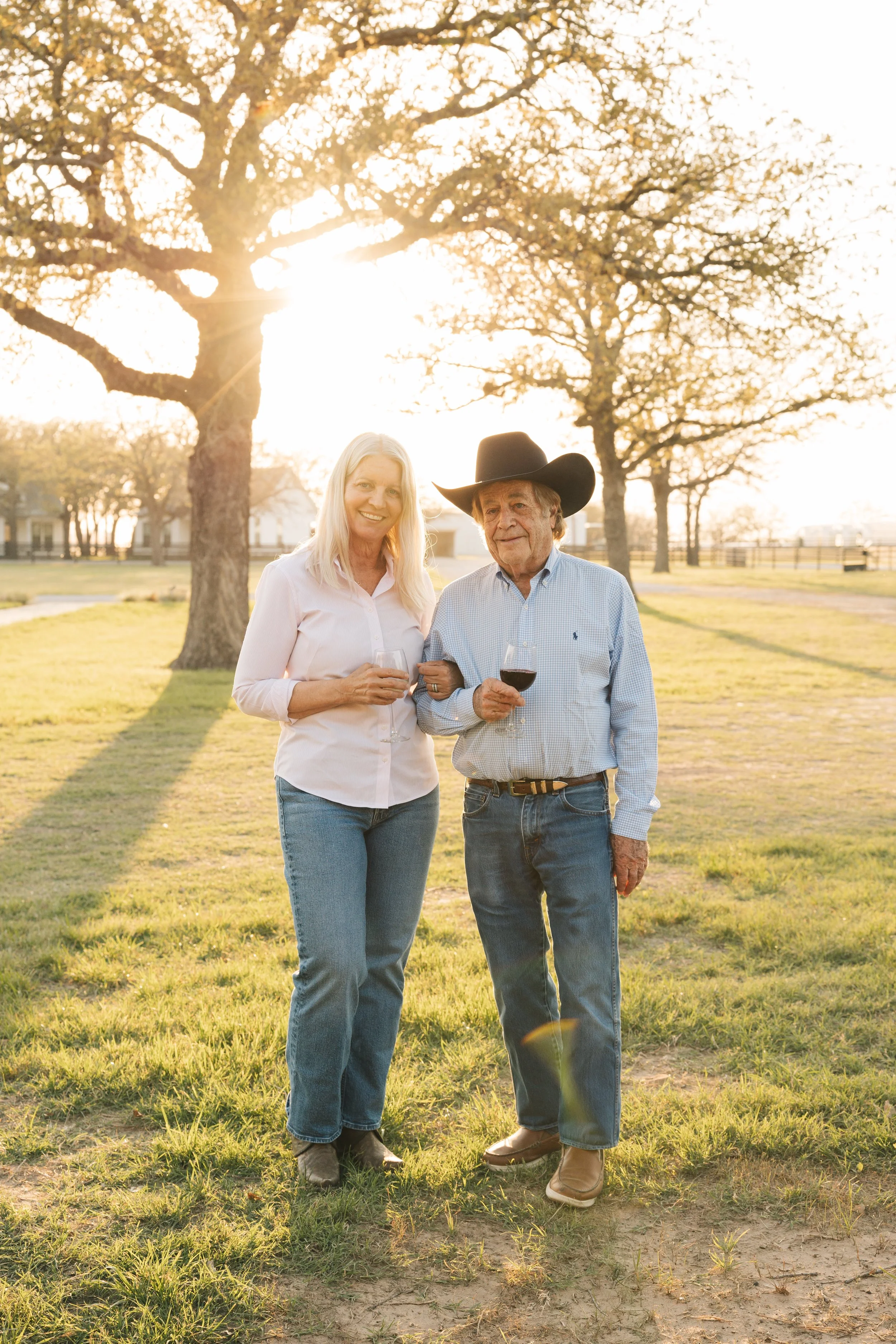 A smiling woman and an older man with a cowboy hat standing outdoors in a park, holding glasses of red wine, with large trees and a setting sun in the background.