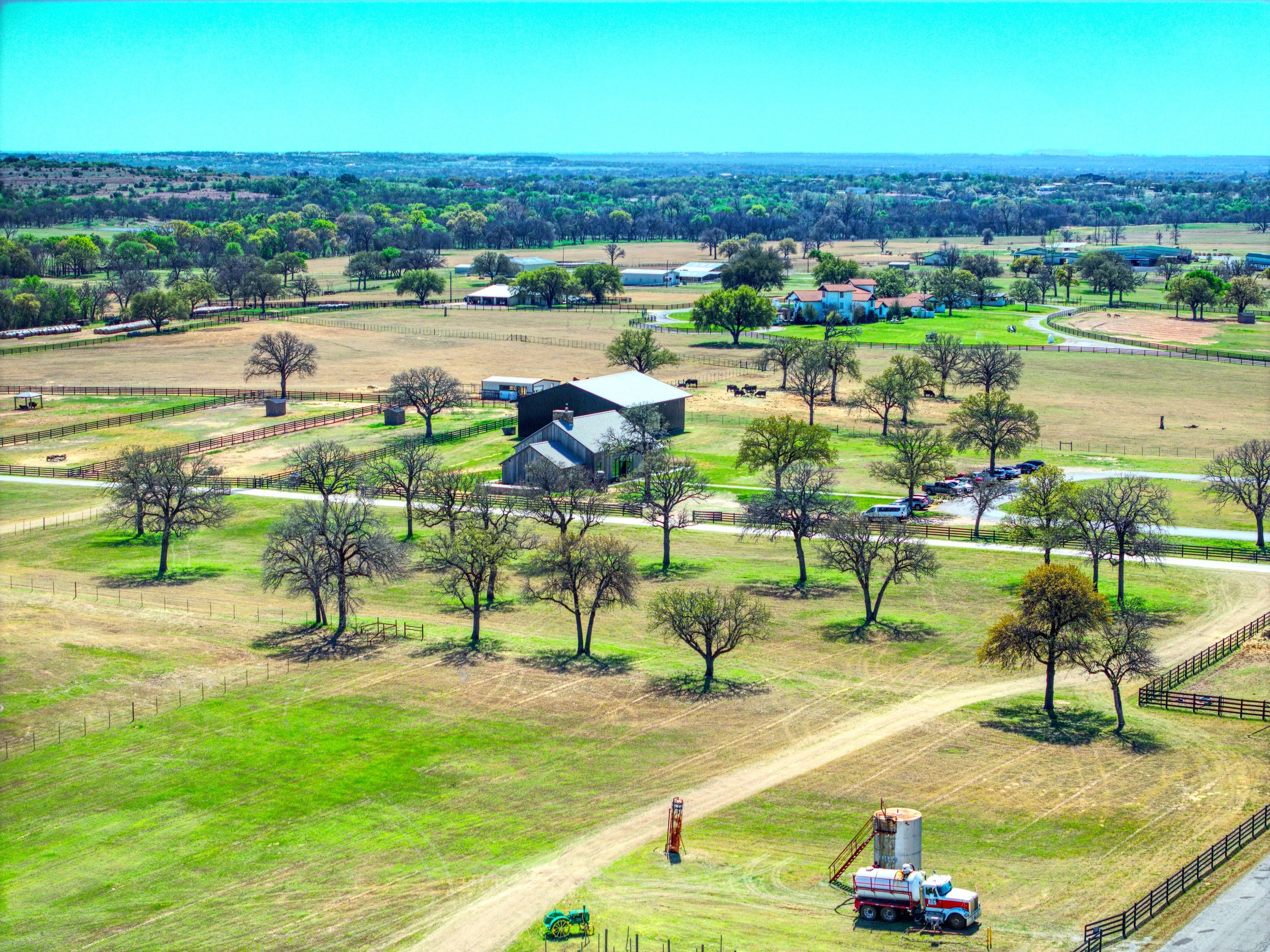 Aerial view of a rural farm landscape with fields, trees, buildings, and vehicles, under a clear blue sky.