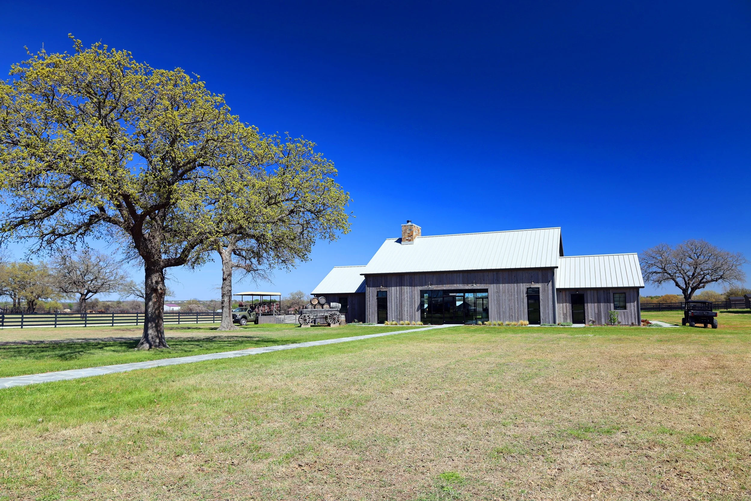 A rural farm scene with a large tree, a barn, and a parking area with golf carts on a sunny day.