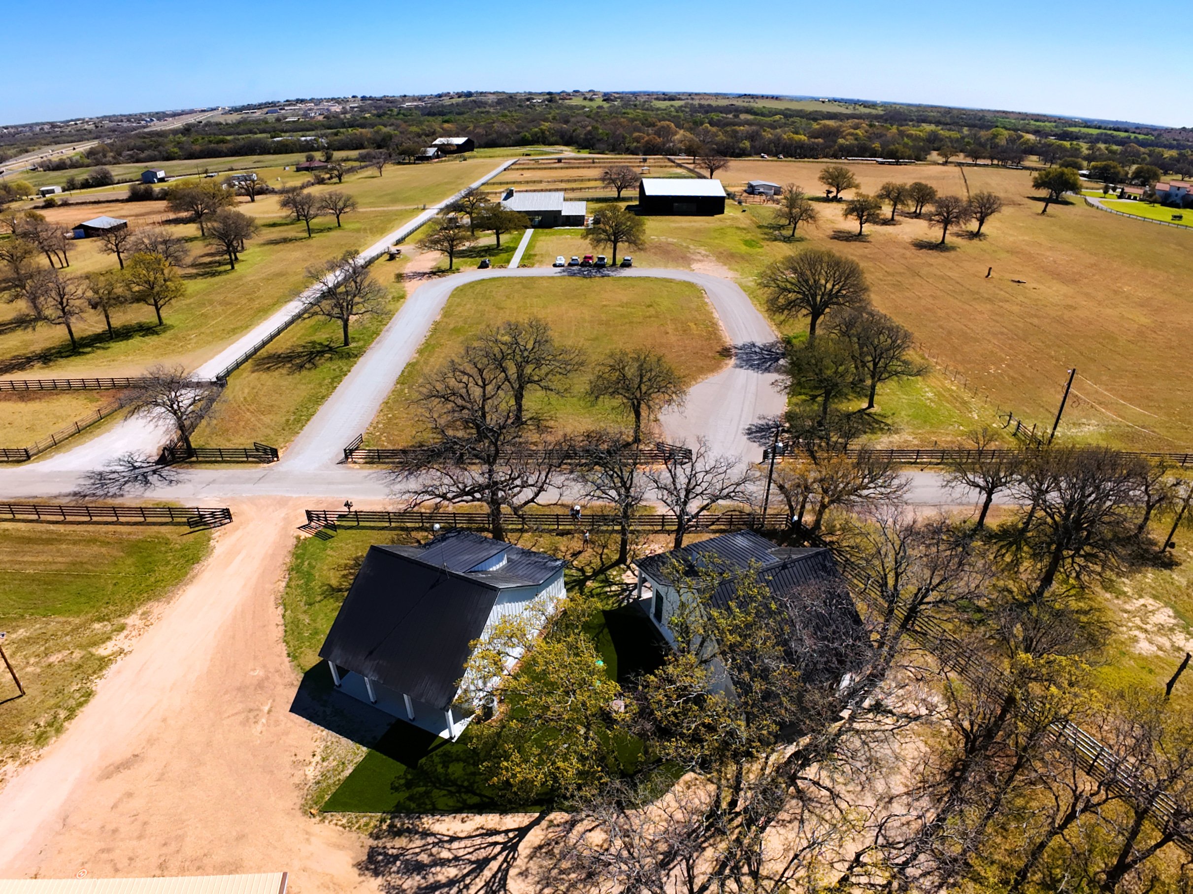 Aerial view of a rural landscape with open fields, scattered trees, driveways, and farm buildings on a sunny day.