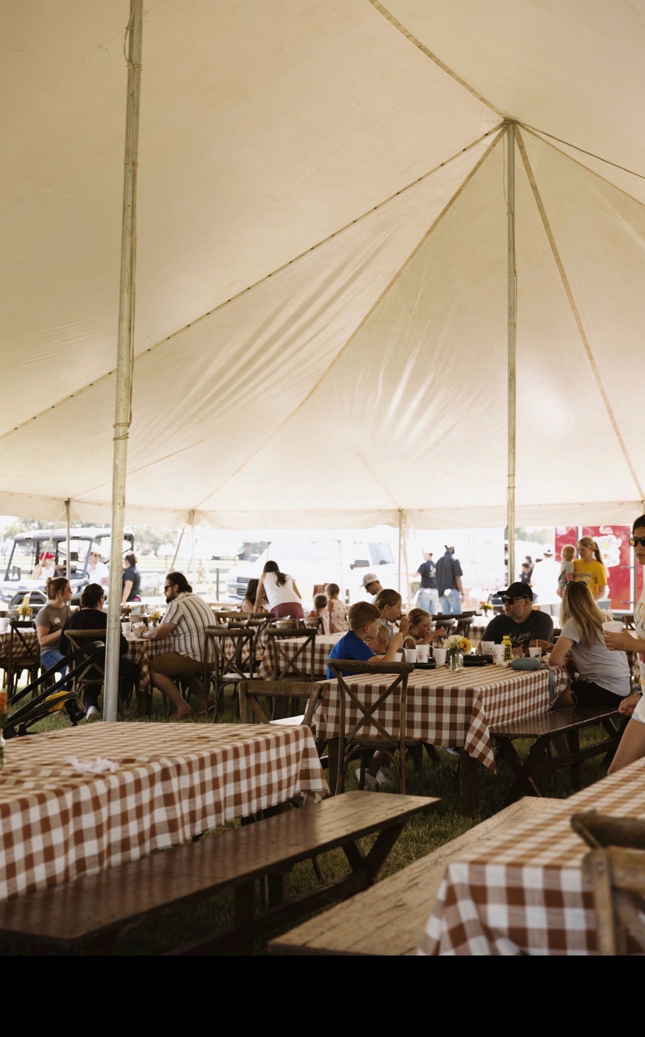 People dining at an outdoor restaurant under a large canopy with checkered tablecloths and wooden picnic tables.