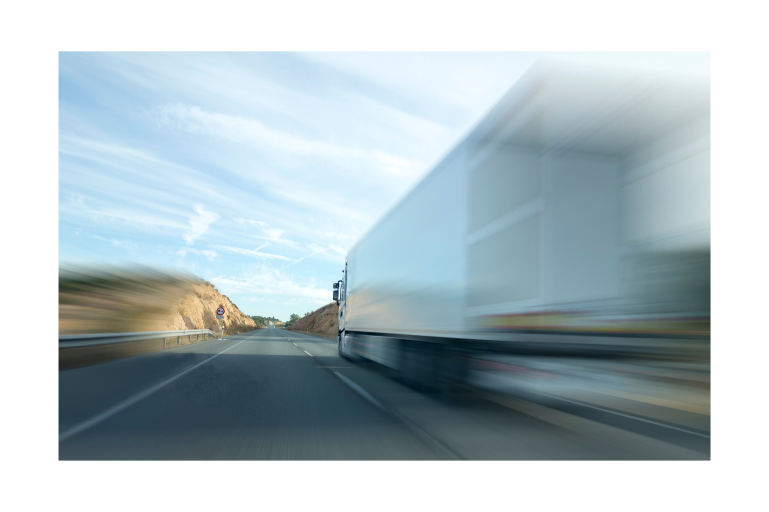 A highway with a large white truck passing by, motion blur effect, blue sky with some clouds, with rocky hills on both sides of the road.