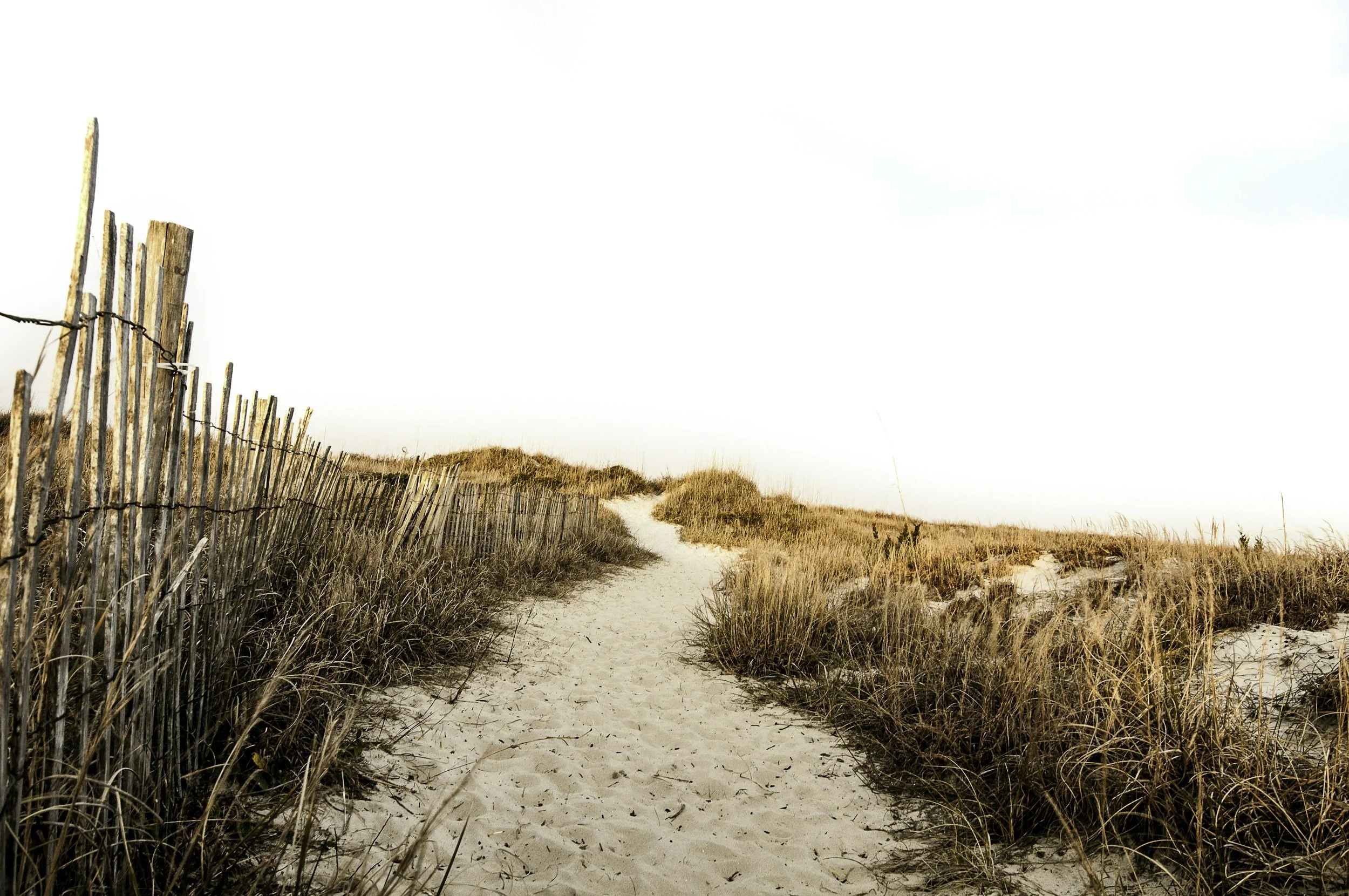 A sandy pathway through dunes with tall grass, bordered by wooden fencing on the left, leading into a clear sky.