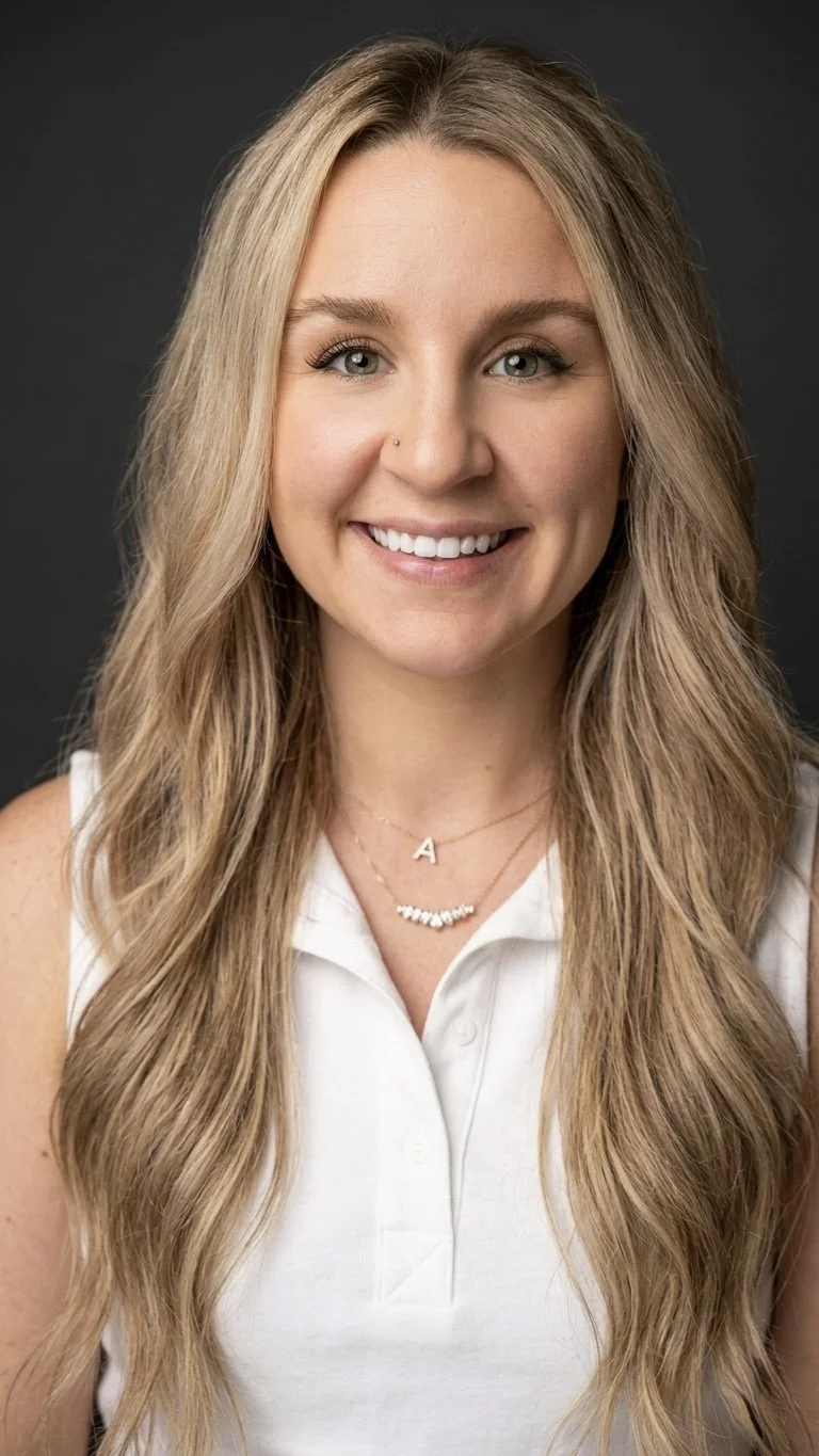 A young woman with long, wavy blonde hair and blue eyes smiling, wearing a white sleeveless top and layered necklaces, one with an 'A' pendant, against a dark gray background.