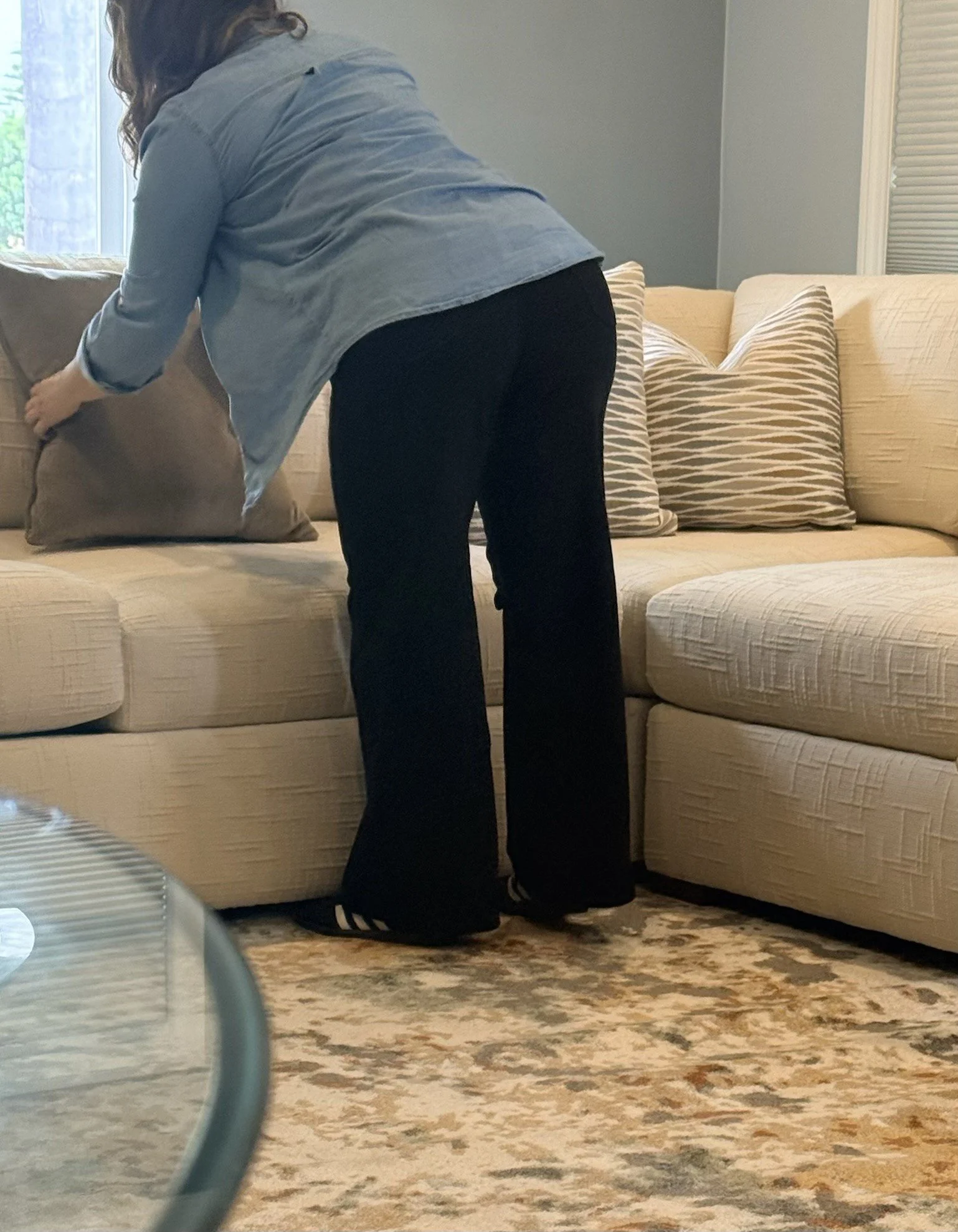 A person with long hair bending down on a beige sofa, with patterned and plain pillows, in a living room with a window and a patterned area rug.