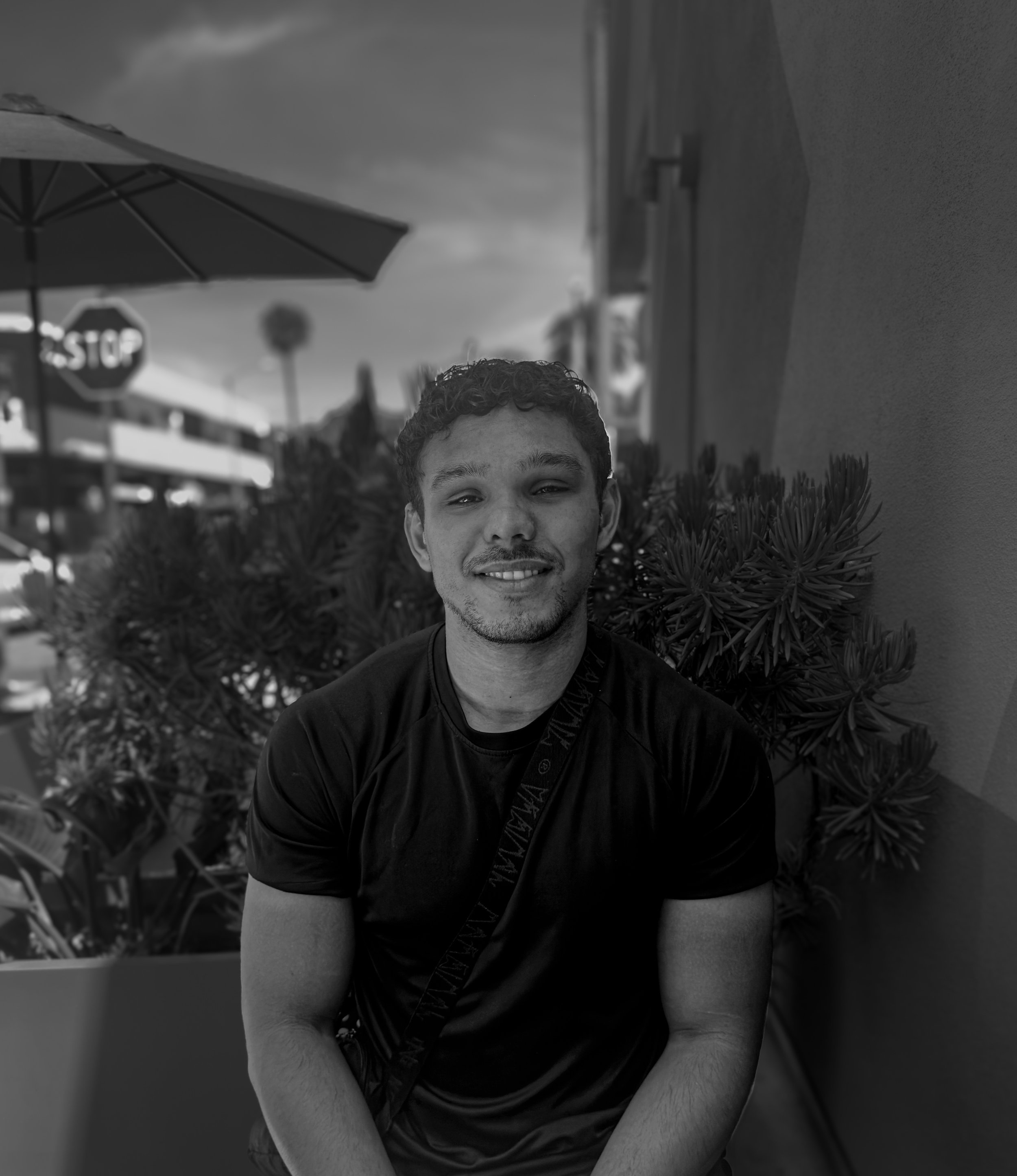 A person with curly hair smiling outdoors, with a stopped sign, an umbrella, and palm trees in the background.