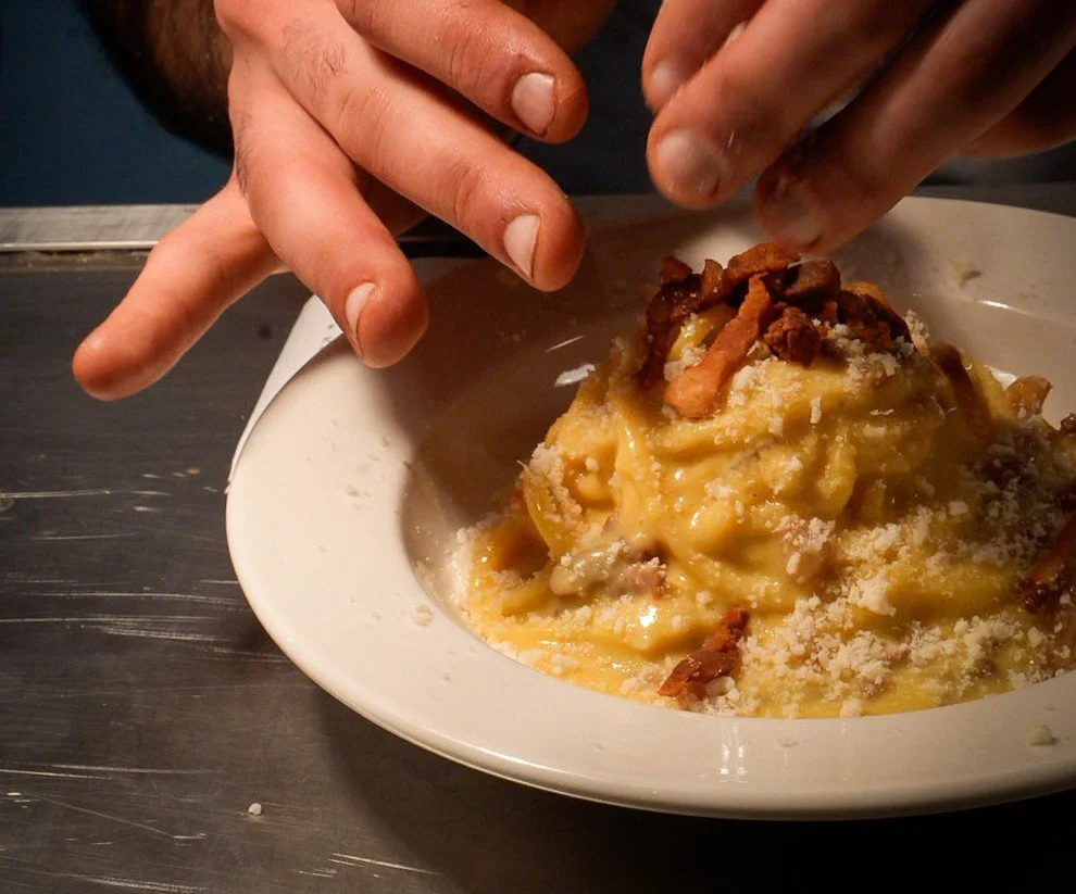 Hands preparing or garnishing a bowl of creamy pasta with bacon bits and grated cheese.
