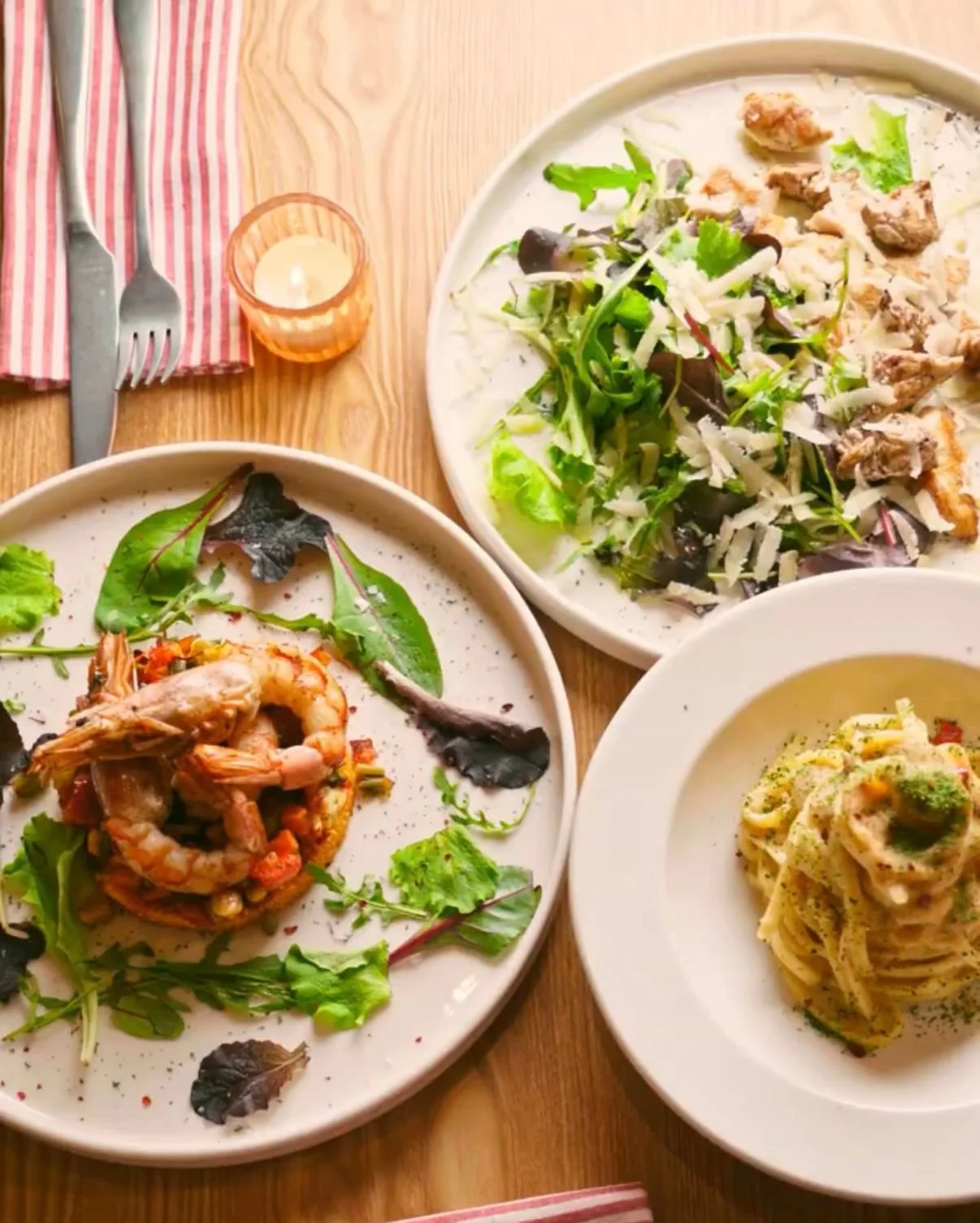 Three plates of food: a seafood dish with shrimp on a bed of vegetables, a mixed greens salad with cheese, and a plate of spaghetti with sauce and herbs. There is a fork with a napkin and a small candle on a wooden table.