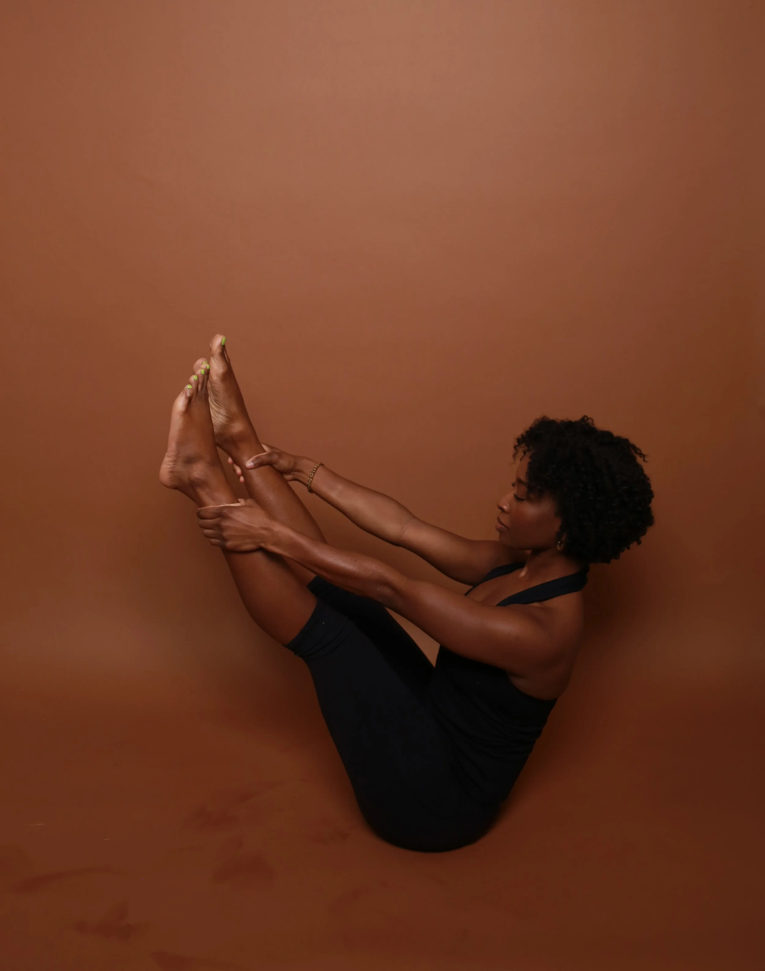 A black Woman in black tank top performing a  pilates open leg rocker pose against a brown background, sitting with her legs held close to her chest and arms extended
