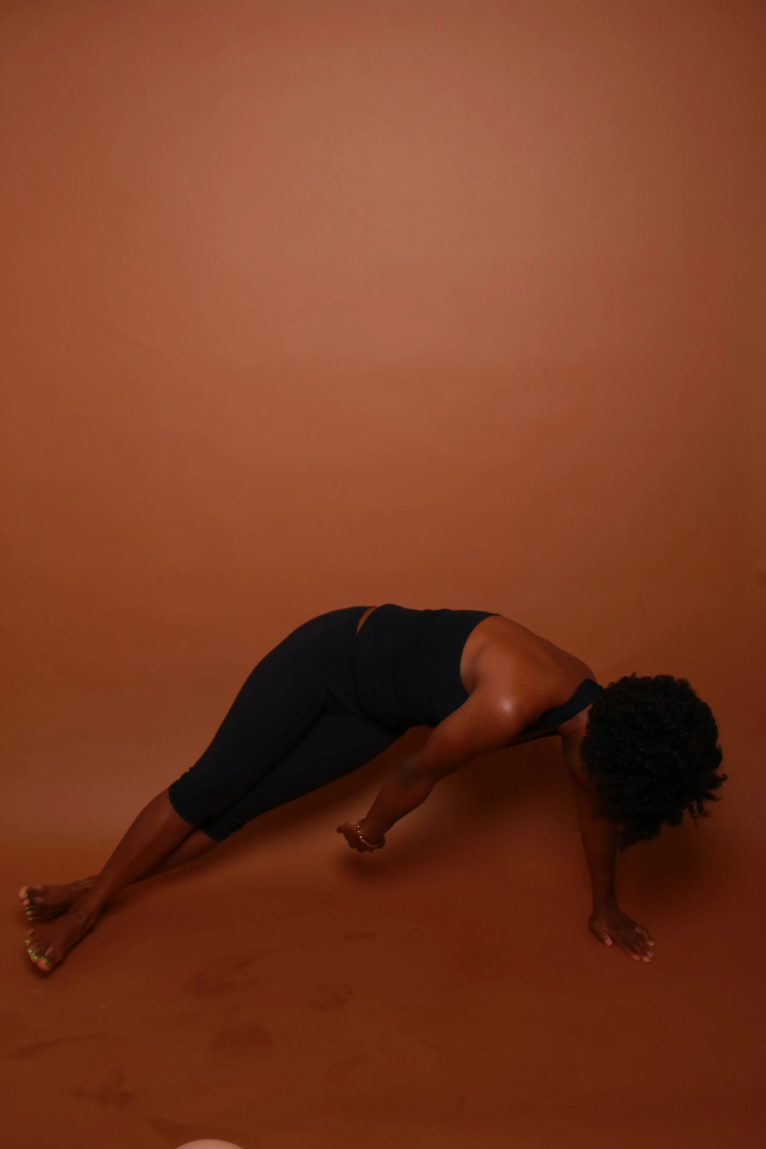 A black woman with curly hair is doing a side plank pilates pose on a brown floor against a matching brown background, wearing black leggings and a black sports bra.