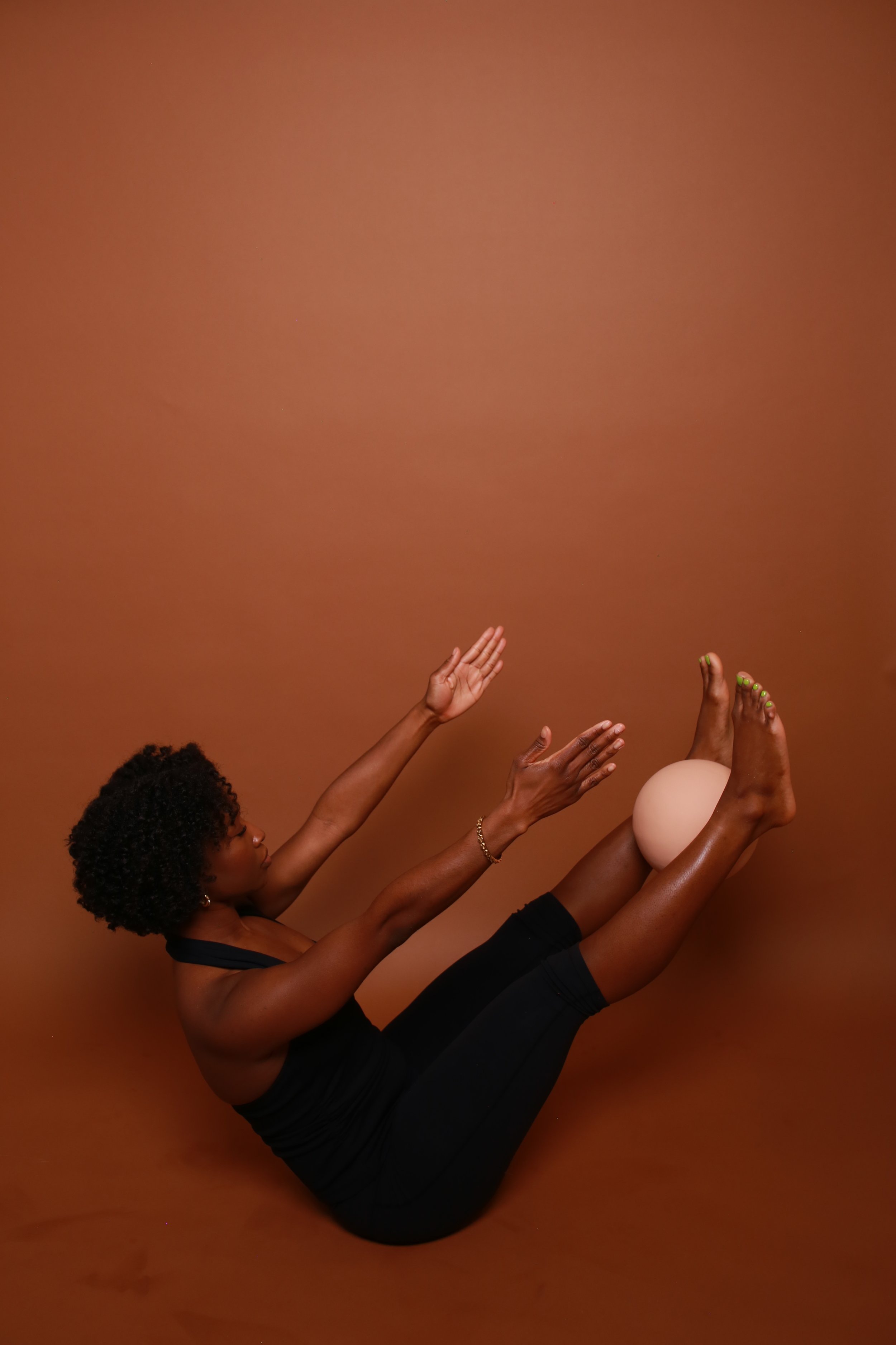 A black woman with curly hair in a black athletic outfit poses against a plain brown background, balancing with her legs raised, holding a Bala exercise ball between her calves, and reaching upward with her arms. Teaser pilates position
