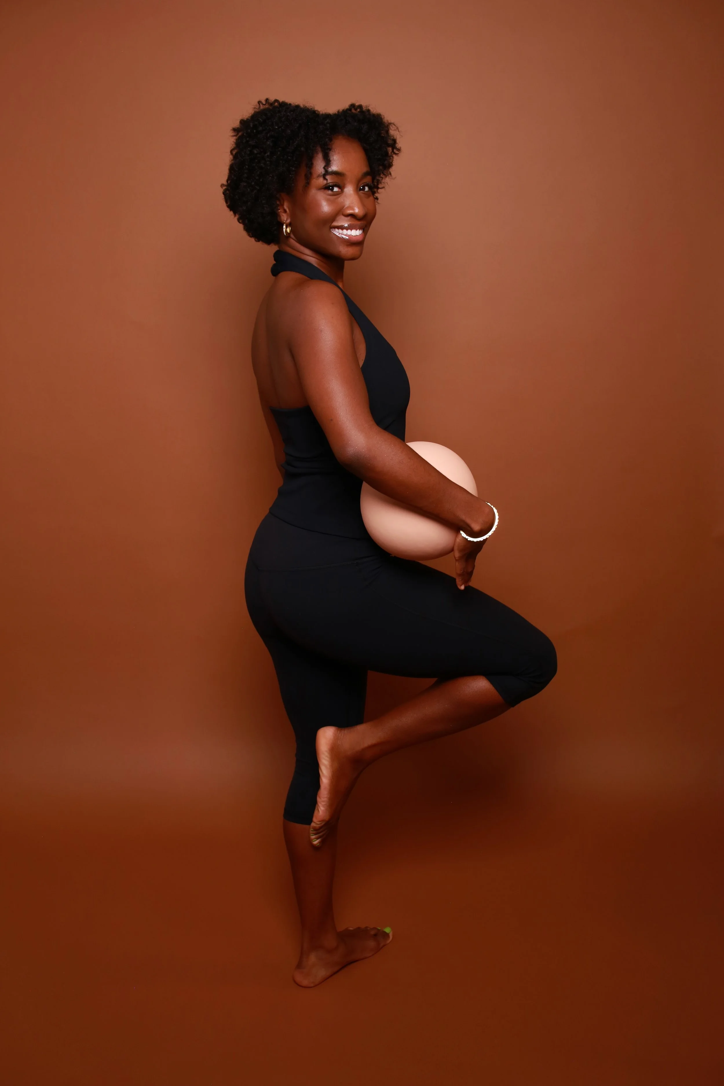 A woman with curly black hair smiling while standing in a yoga pose against a brown background, holding a beige exercise ball, wearing a black tank top and black leggings.
