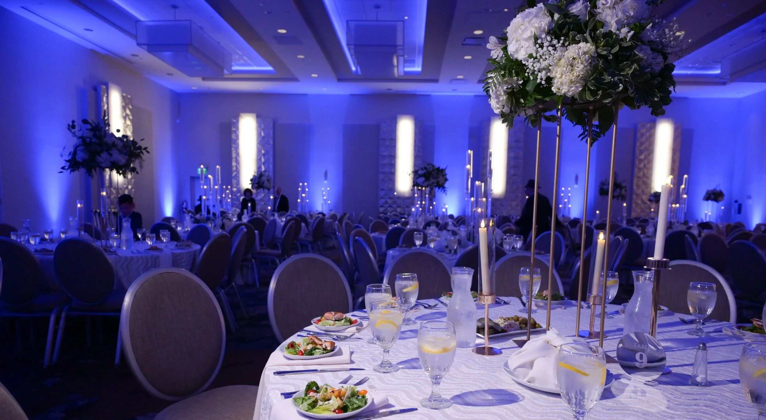 Elegant banquet hall decorated for a formal event with round tables, floral centerpieces, candles, and dim blue lighting.