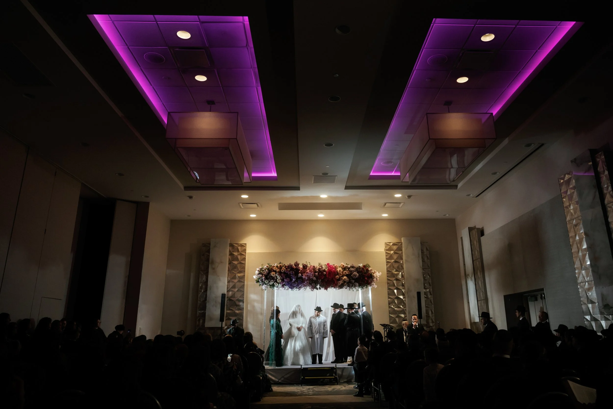 A wedding ceremony taking place indoors with a bride and groom standing together in front of an officiant, surrounded by guests. The ceiling features purple lighting accents and there is a large floral arrangement in front of the couple.