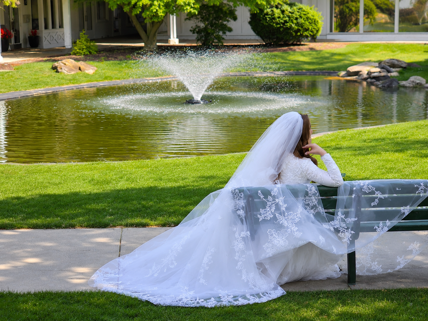 A bride sitting on a park bench near a pond with a fountain, wearing a white lace wedding gown and veil, resting her chin on her hand, and looking away from the camera.
