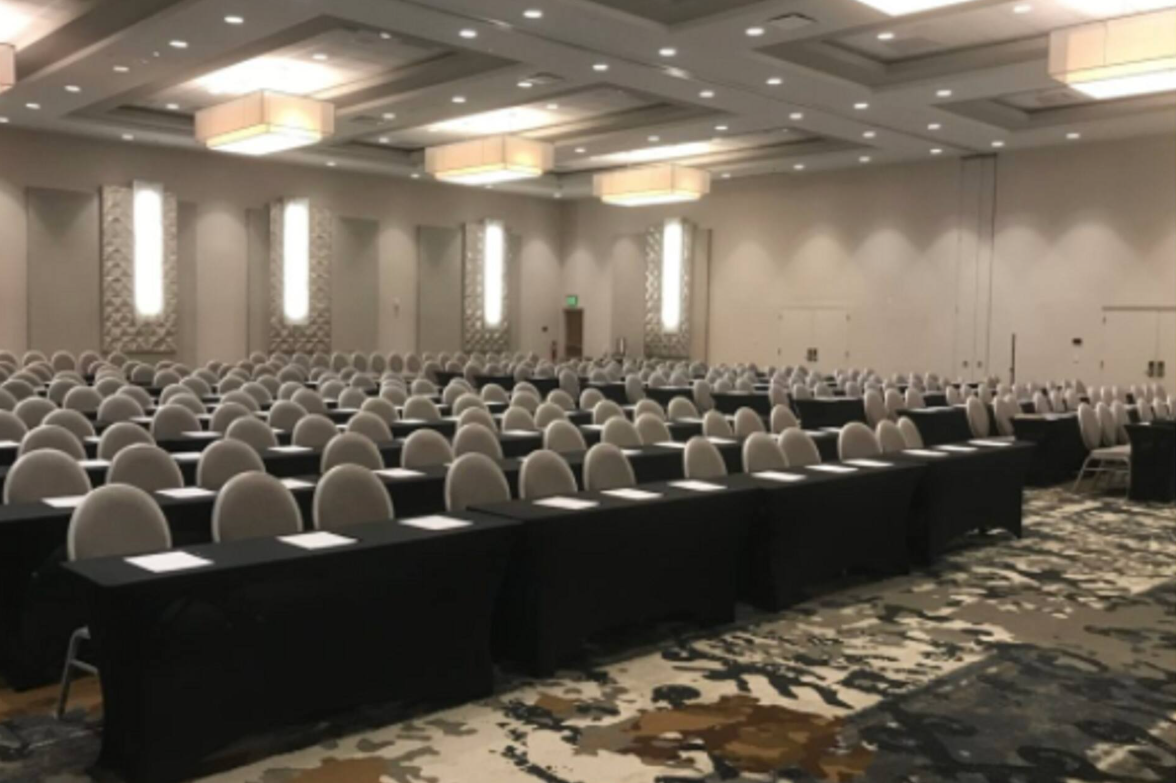 Conference room with long tables covered in black cloth and white chairs arranged in rows, with name cards on the tables, in a well-lit, spacious setting.