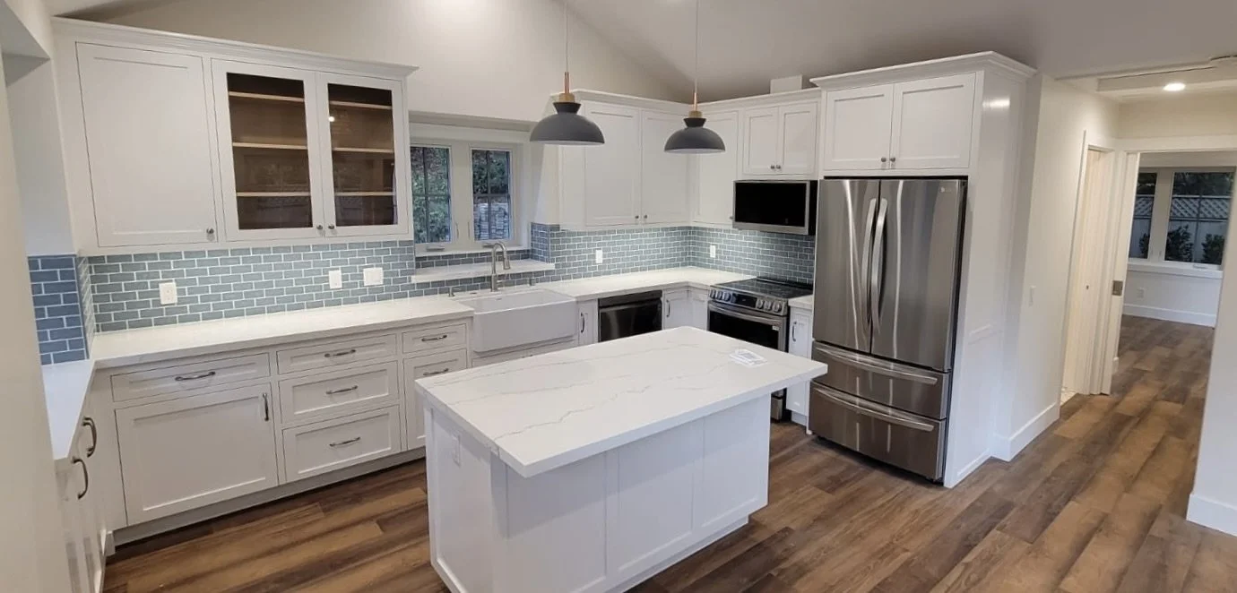 Modern kitchen with white cabinets, stainless steel refrigerator, black stove, blue tile backsplash, and a central island with a white countertop. There are pendant lights above the island, and a window above the sink.