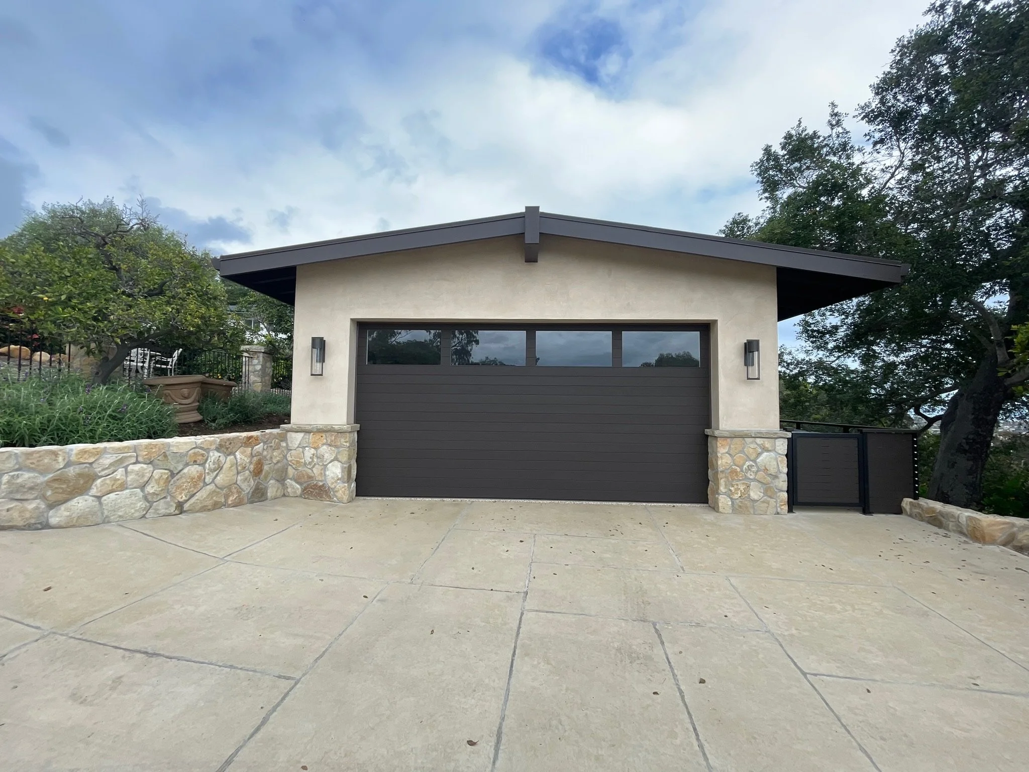 A modern house with a flat roof, a large garage door, surrounded by greenery and trees, with a stone wall in the front, and a concrete patio in the foreground.