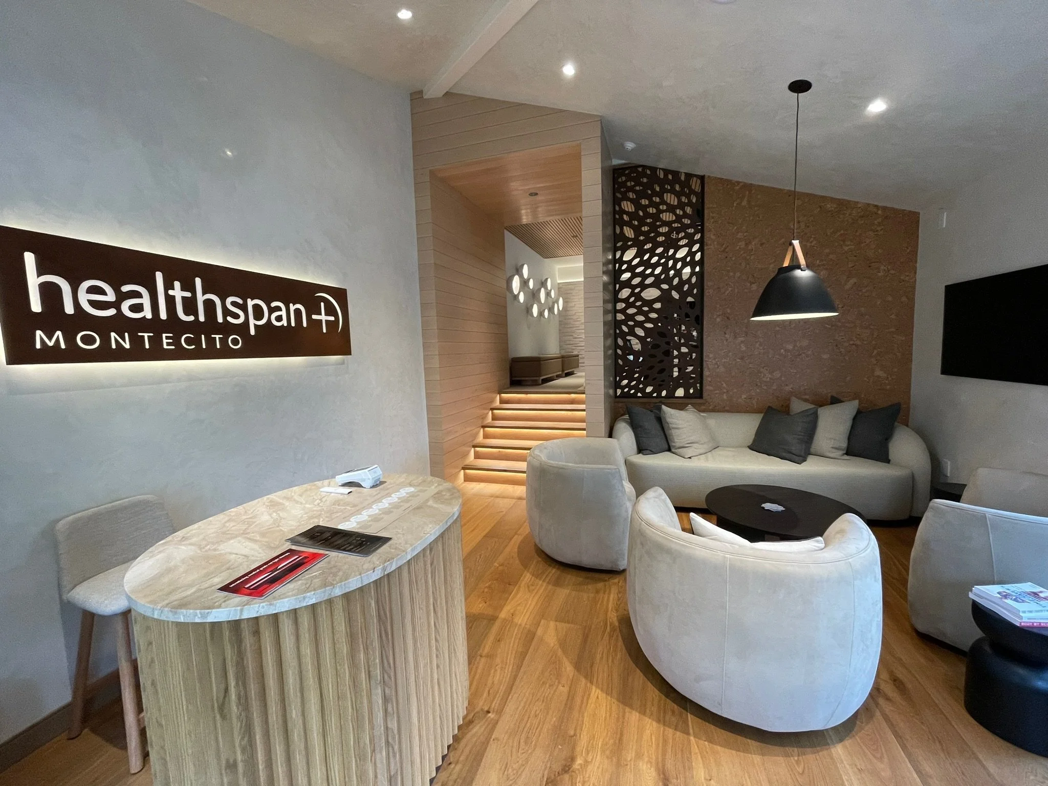 Interior of a modern medical or healthcare facility with a sign reading 'healthspan+ Montecito,' a curved reception desk, cream-colored seating, and decorative wooden and dark metal room dividers.
