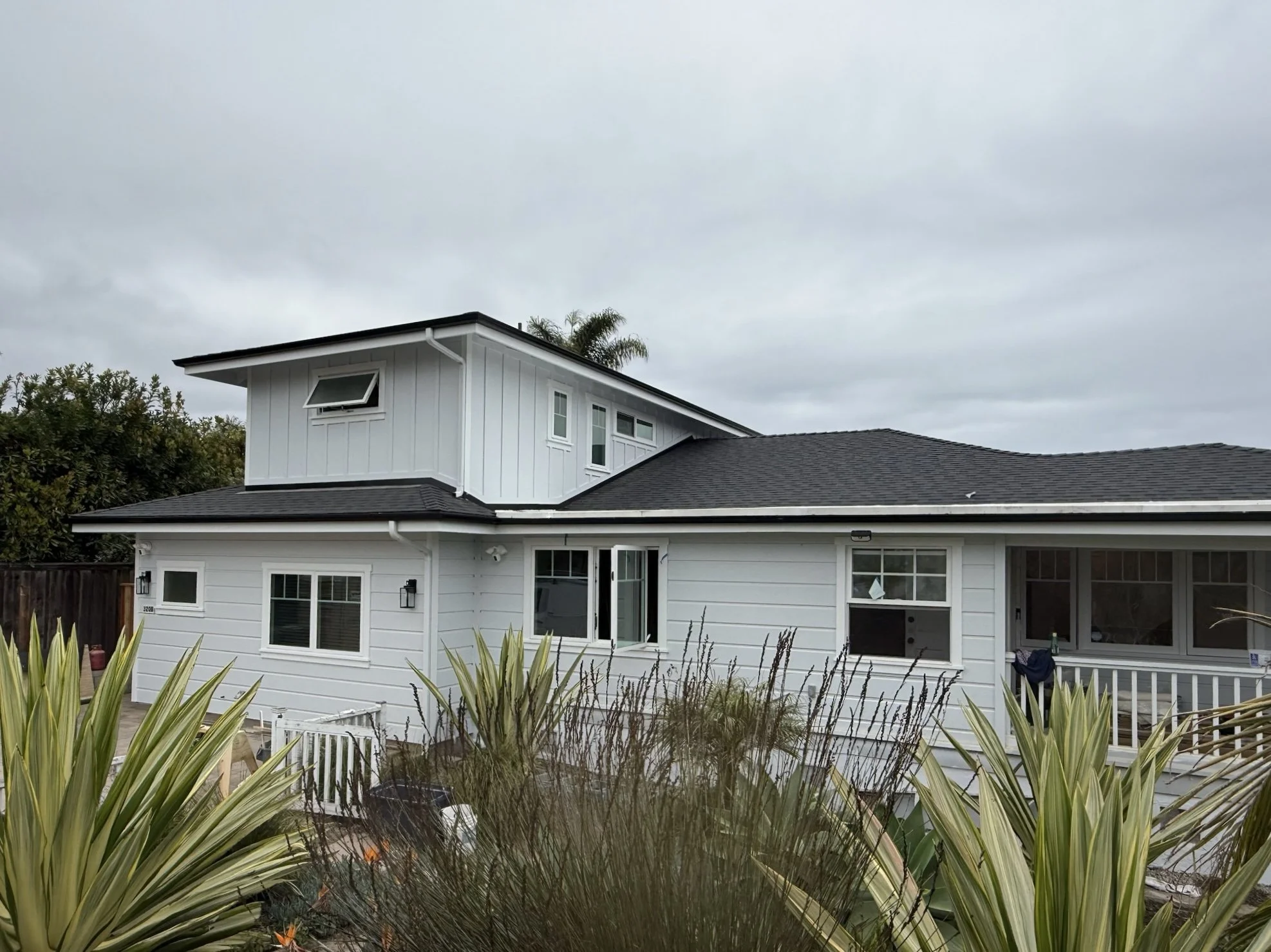 A two-story white house with a black roof, multiple windows, and a screened porch under cloudy skies, surrounded by green plants and trees.