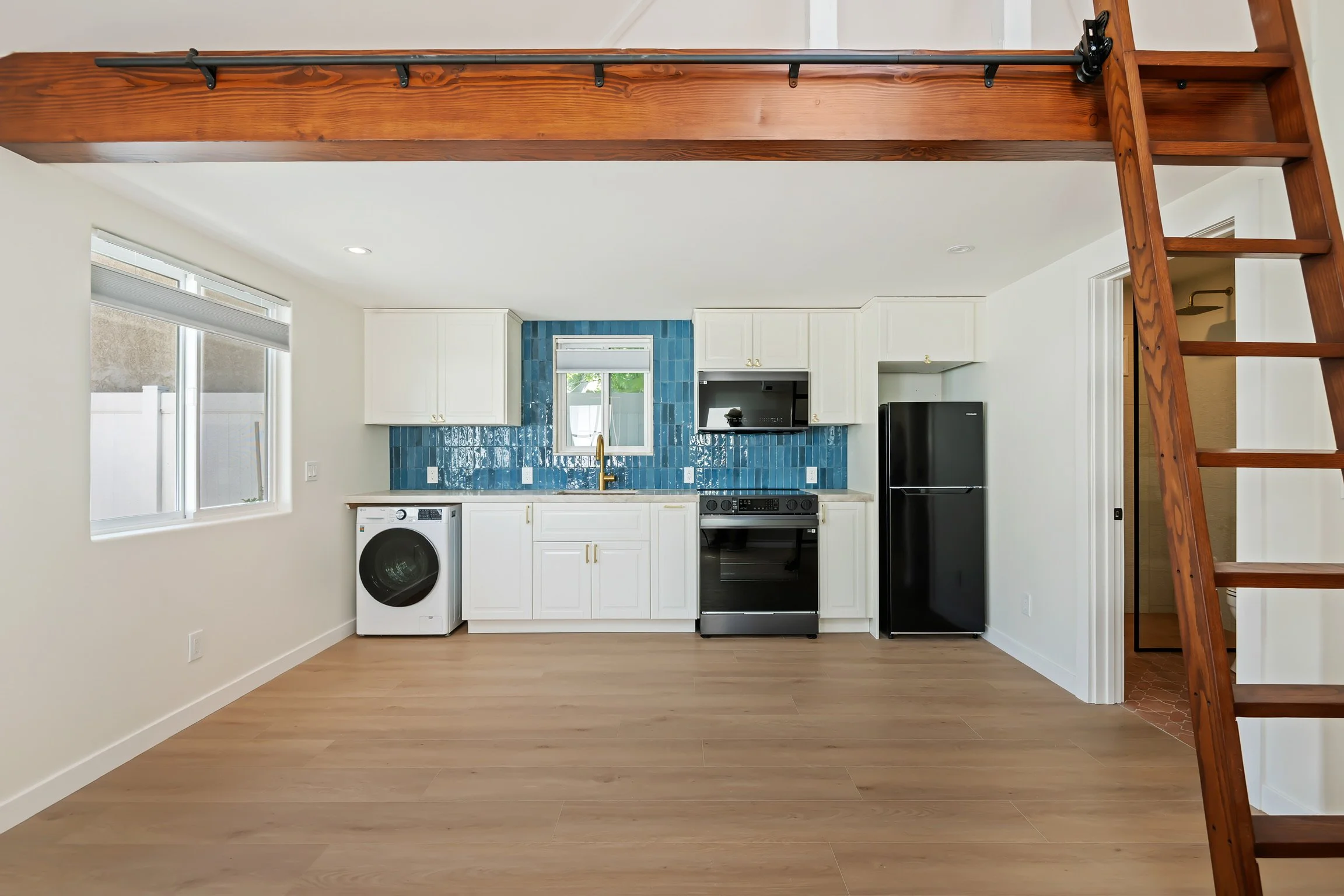 Interior view of a modern kitchen with white cabinets, blue tiled backsplash, black appliances, and a wooden loft ladder.