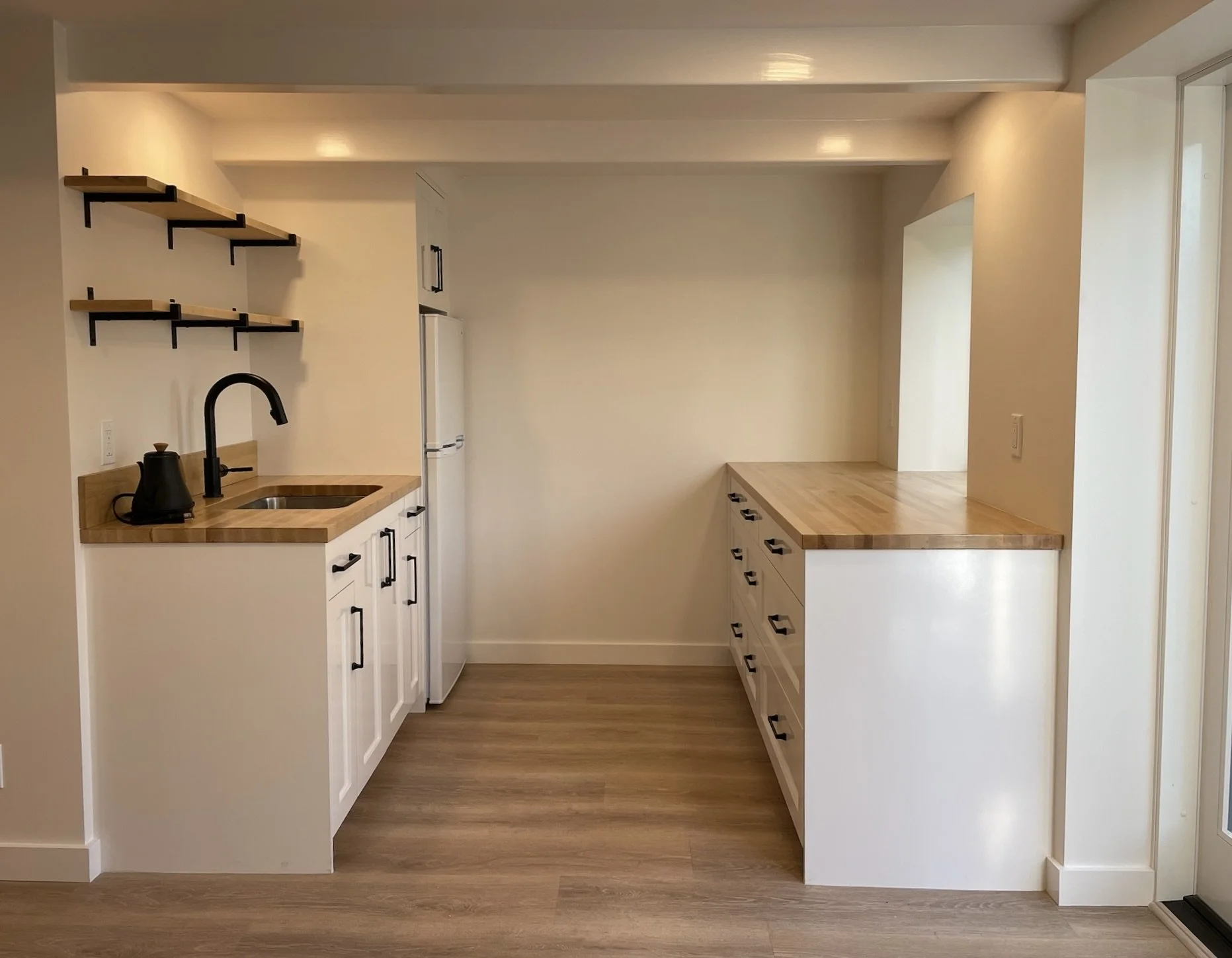 A small kitchen with white cabinets, wood countertops, and a black faucet. Open shelving on the left wall and a side-by-side refrigerator in the corner. The kitchen island has drawers on the right side. The floor is wood, and the walls are painted a light color.