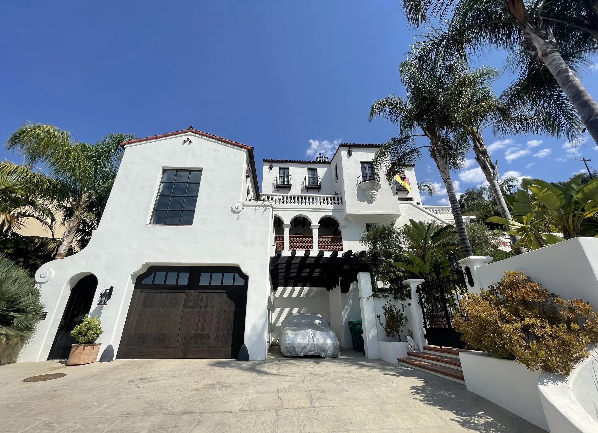 A large white house with arched windows and balconies, surrounded by palm trees and plants, under a blue sky with some clouds.