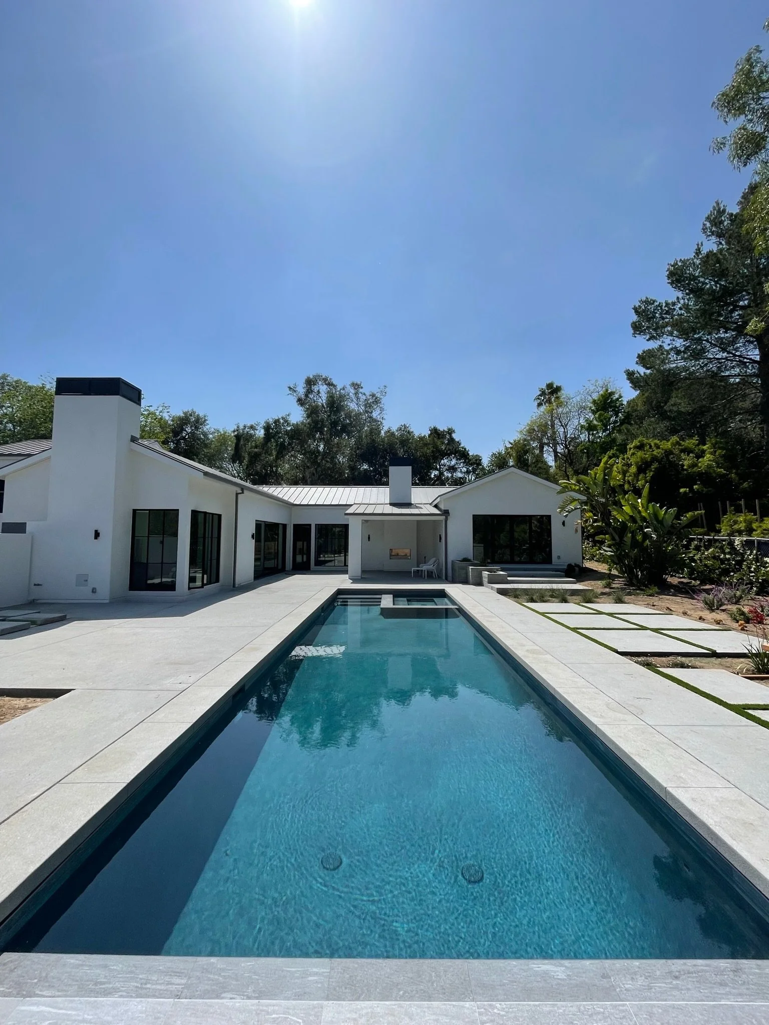 Modern white house with large windows and a rectangular swimming pool in the backyard, surrounded by trees and clear blue sky.
