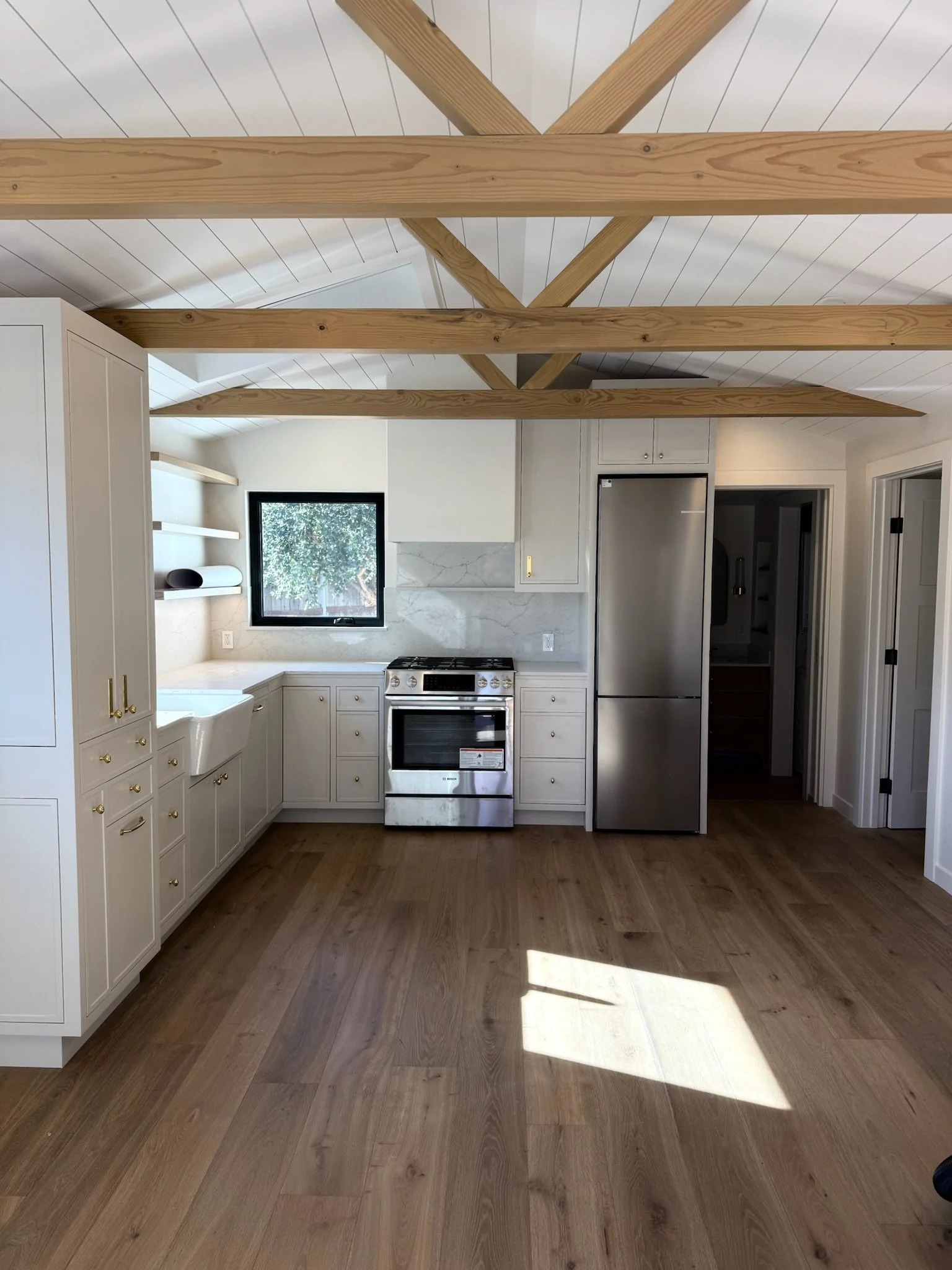 A modern kitchen with white cabinets, a stainless steel stove, a refrigerator, and wooden ceiling beams. There is a window above the sink and a door to another room on the right.