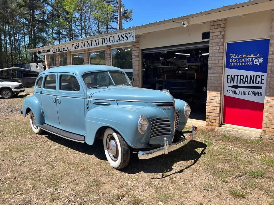 A vintage light blue sedan parked outside an auto glass shop.