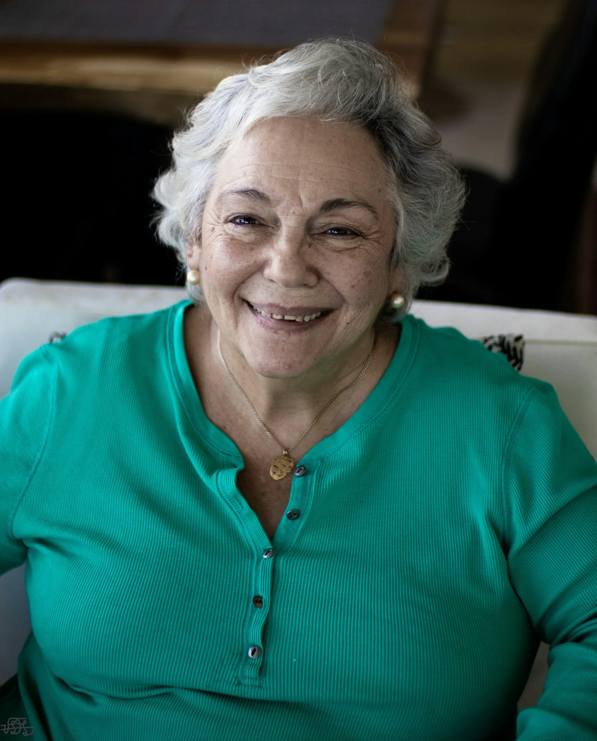 Close-up of an elderly woman with short gray hair smiling, wearing a teal shirt, pearl earrings, a necklace with a gold pendant, and a small charm attached to her shirt.