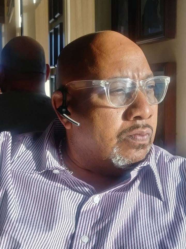 A man wearing clear eyeglasses and a striped button-up shirt, sitting indoors with sunlight on his face, wearing a Bluetooth headset.