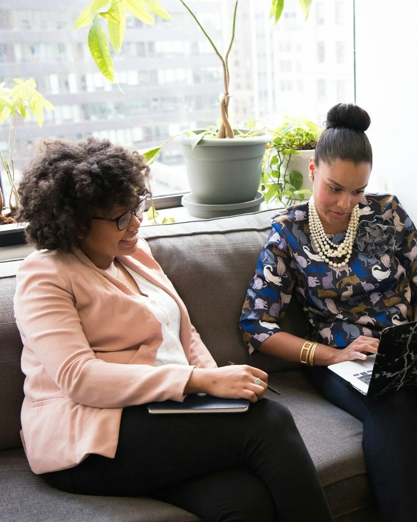 Two women are sitting on a gray sofa in an office, looking at a laptop. One woman has curly black hair, glasses, and is wearing a light pink blazer and white blouse. The other woman has dark hair in a bun, wearing a blue dress with white swans and pearl necklaces. There are potted plants on the windowsill behind them.