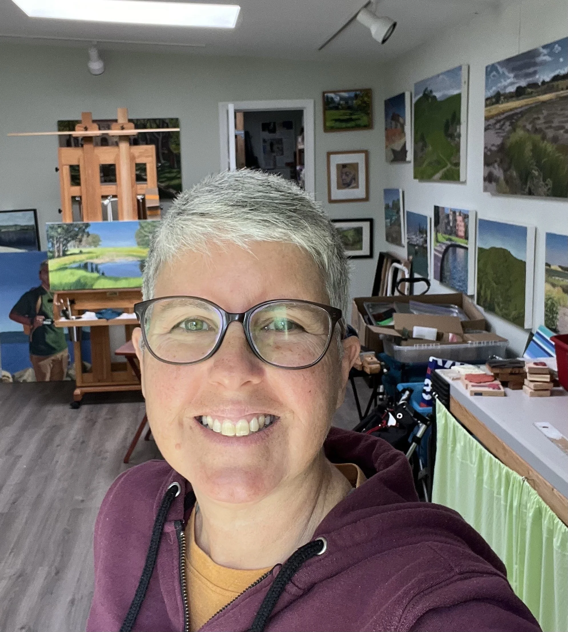 A woman with short gray hair, wearing glasses and a maroon hoodie, smiling and taking a selfie in an art studio filled with landscape paintings on the walls.