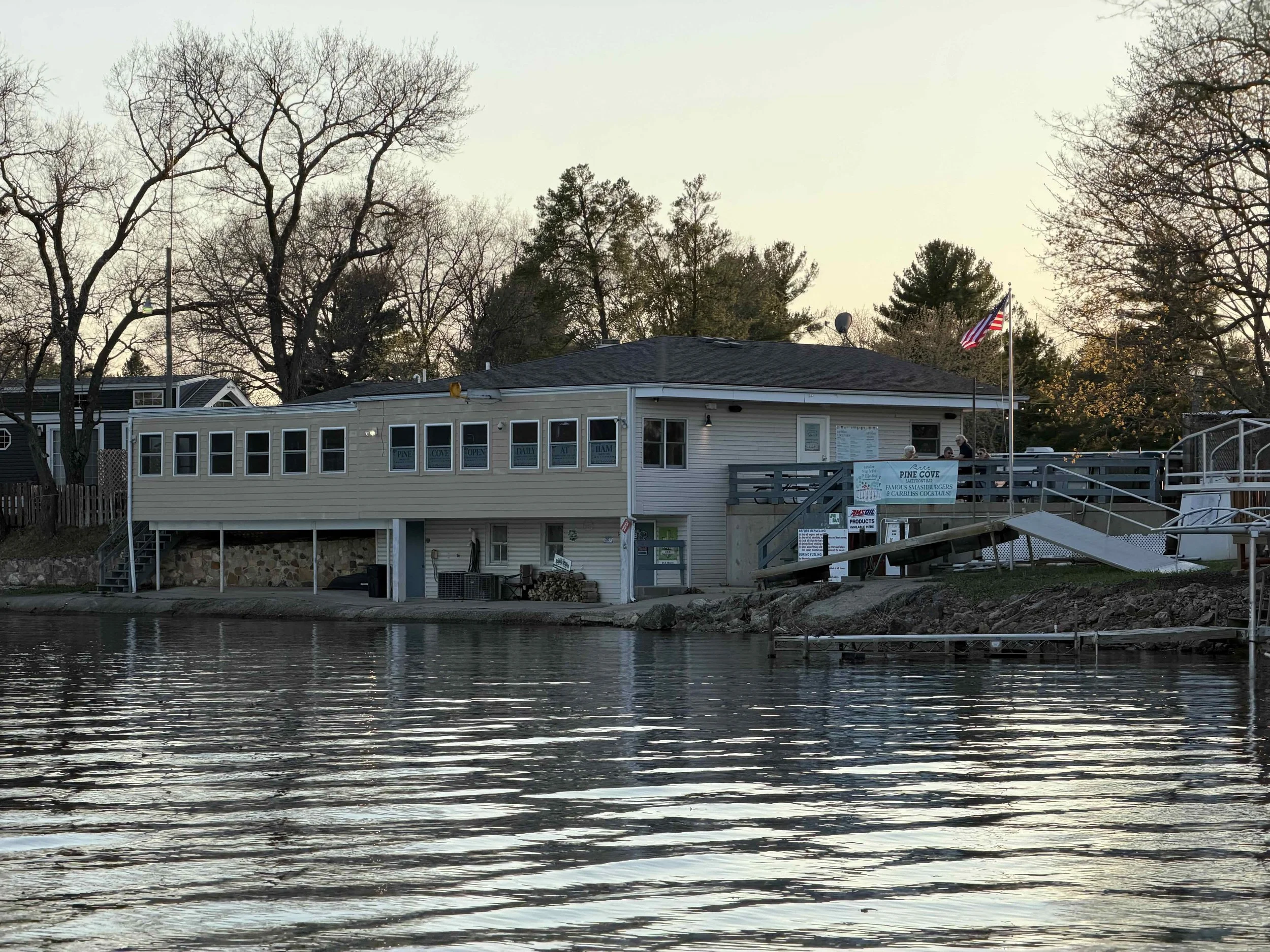 A building by a body of water with leafless trees in the background and an American flag flying on a pole.
