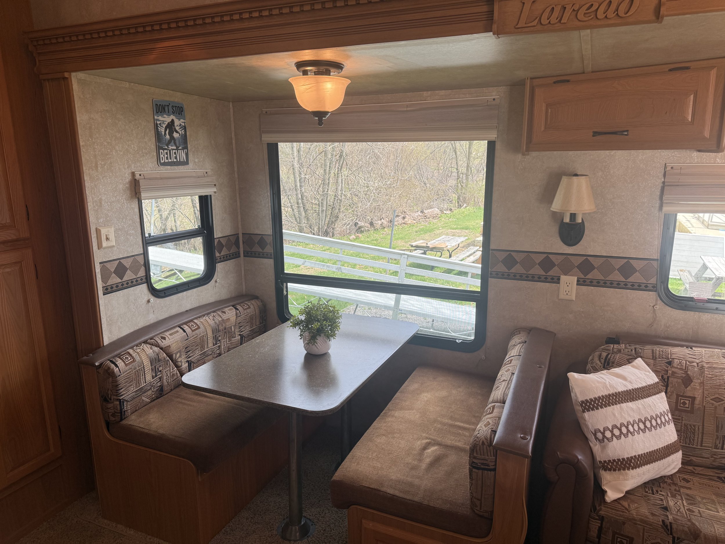 Interior of a camper or RV with a wooden cabinet, a table with a potted plant, and cushioned seating with patterned upholstery. Large windows show an outdoor view of trees and grass.
