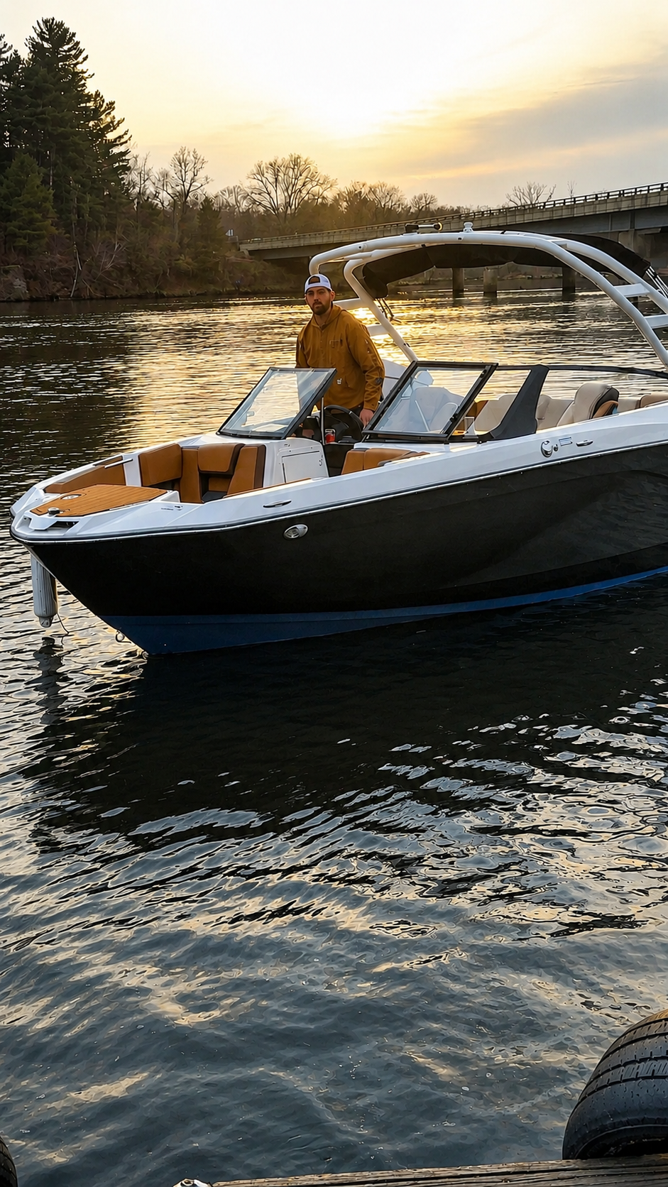 A man in a yellow jacket and white cap standing at the helm of a black and white motorboat on a lake during sunset, with trees and a bridge in the background.