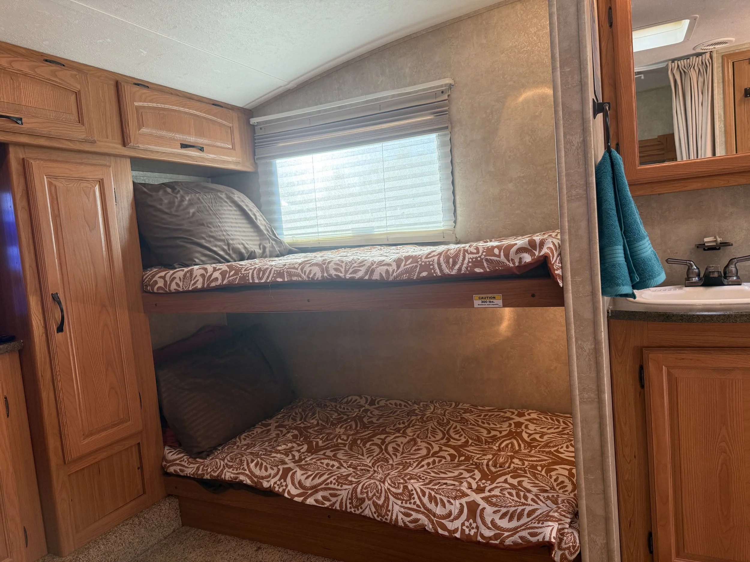 The interior of a recreational vehicle bathroom with a bunk bed area above, featuring wooden cabinetry, a window with blinds, and a beige countertop with teal towels.