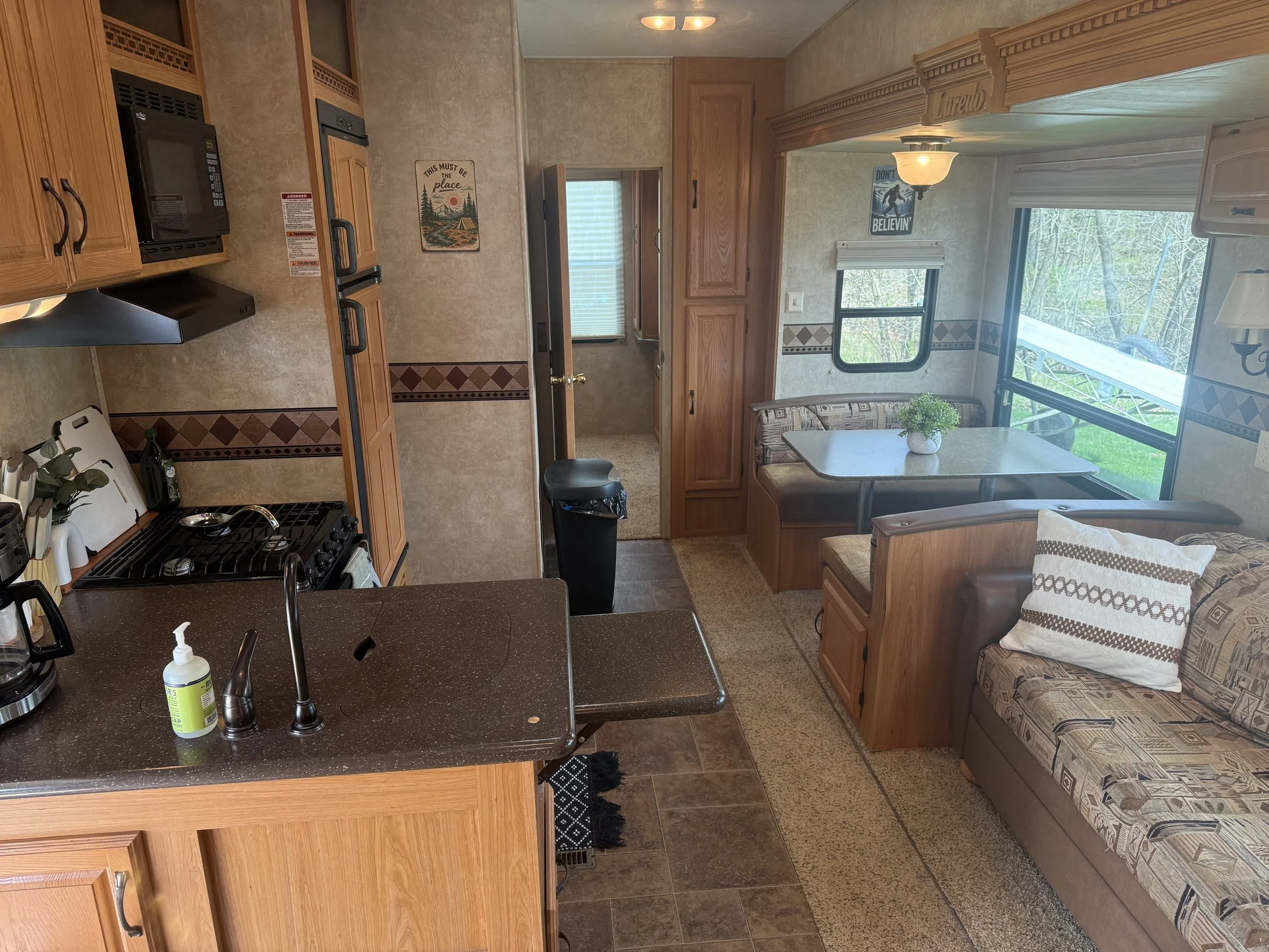 Inside a compact camper or RV, showing a kitchen with a black countertop, stove, and microwave, a dining area with a table and window, and a small hallway leading to a bedroom with a window and a couch.