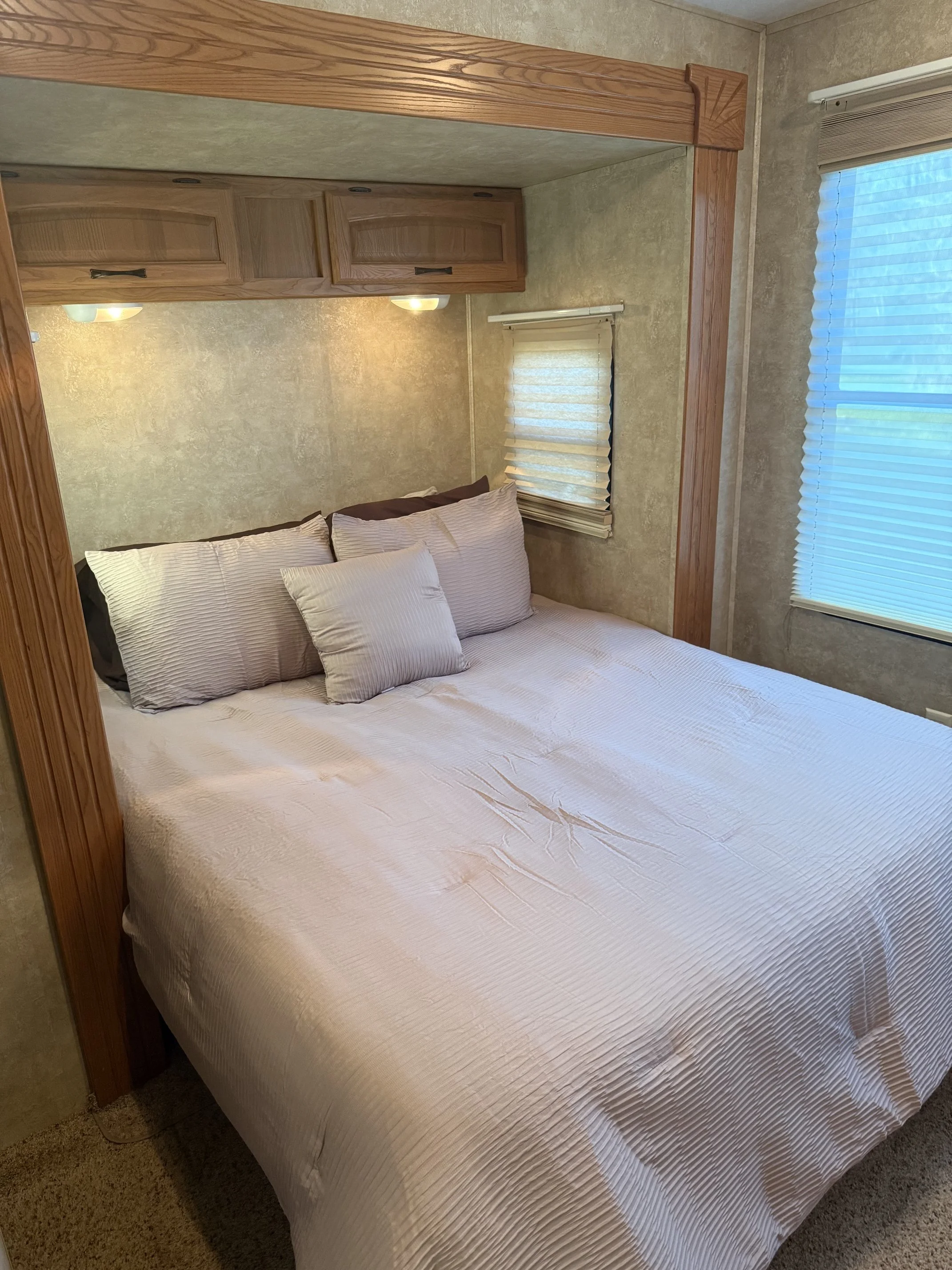 A neatly made bed with white bedspread and two matching pillows in a room with beige textured walls, two windows with blinds, and wooden trim around a mirror frame.
